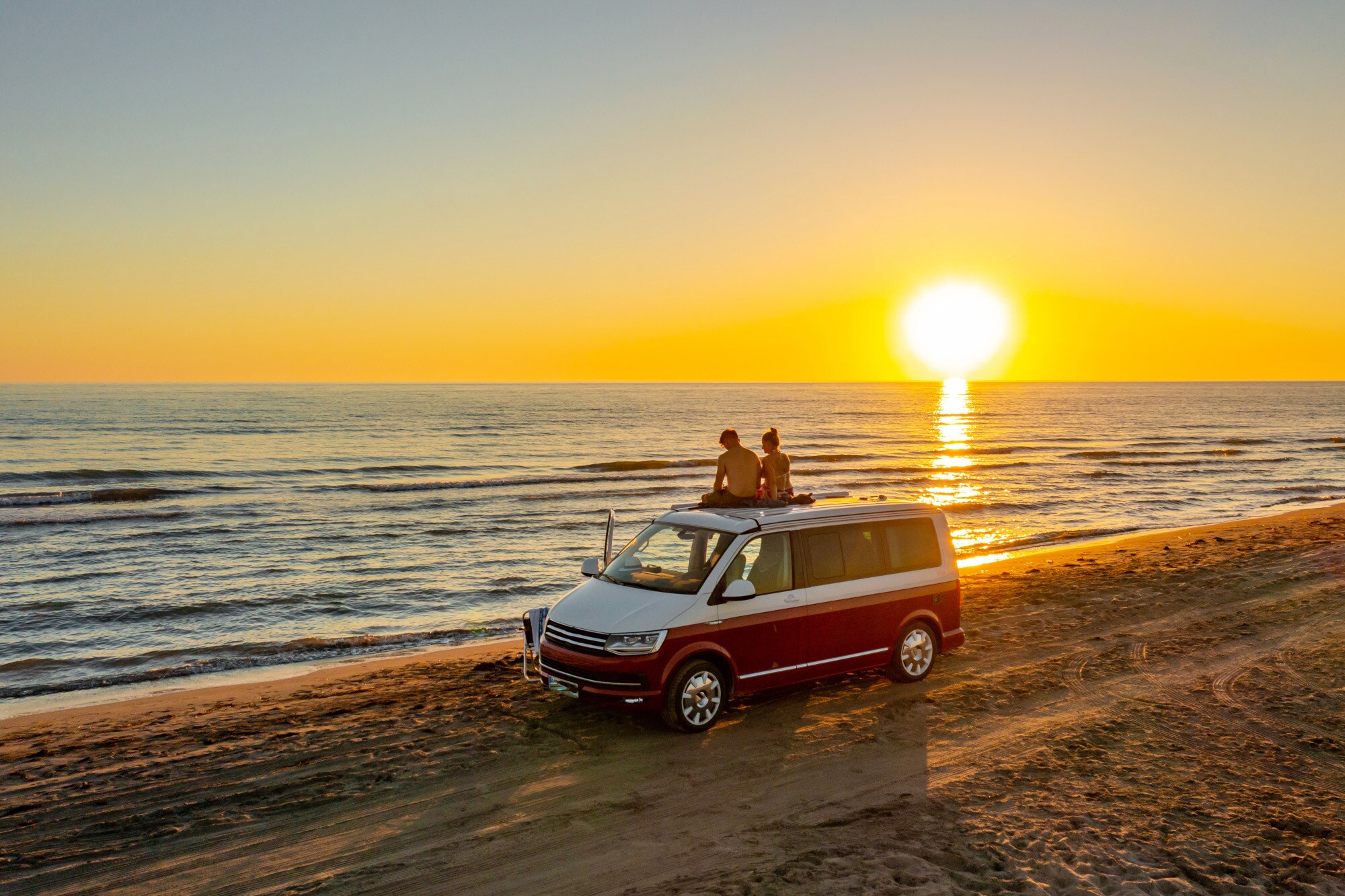 Rückansicht eines Paares auf dem Dach eines Vans am Strand bei Sonnenuntergang. Rückansicht eines Paares auf dem Dach eines Vans am Strand bei Sonnenuntergang.