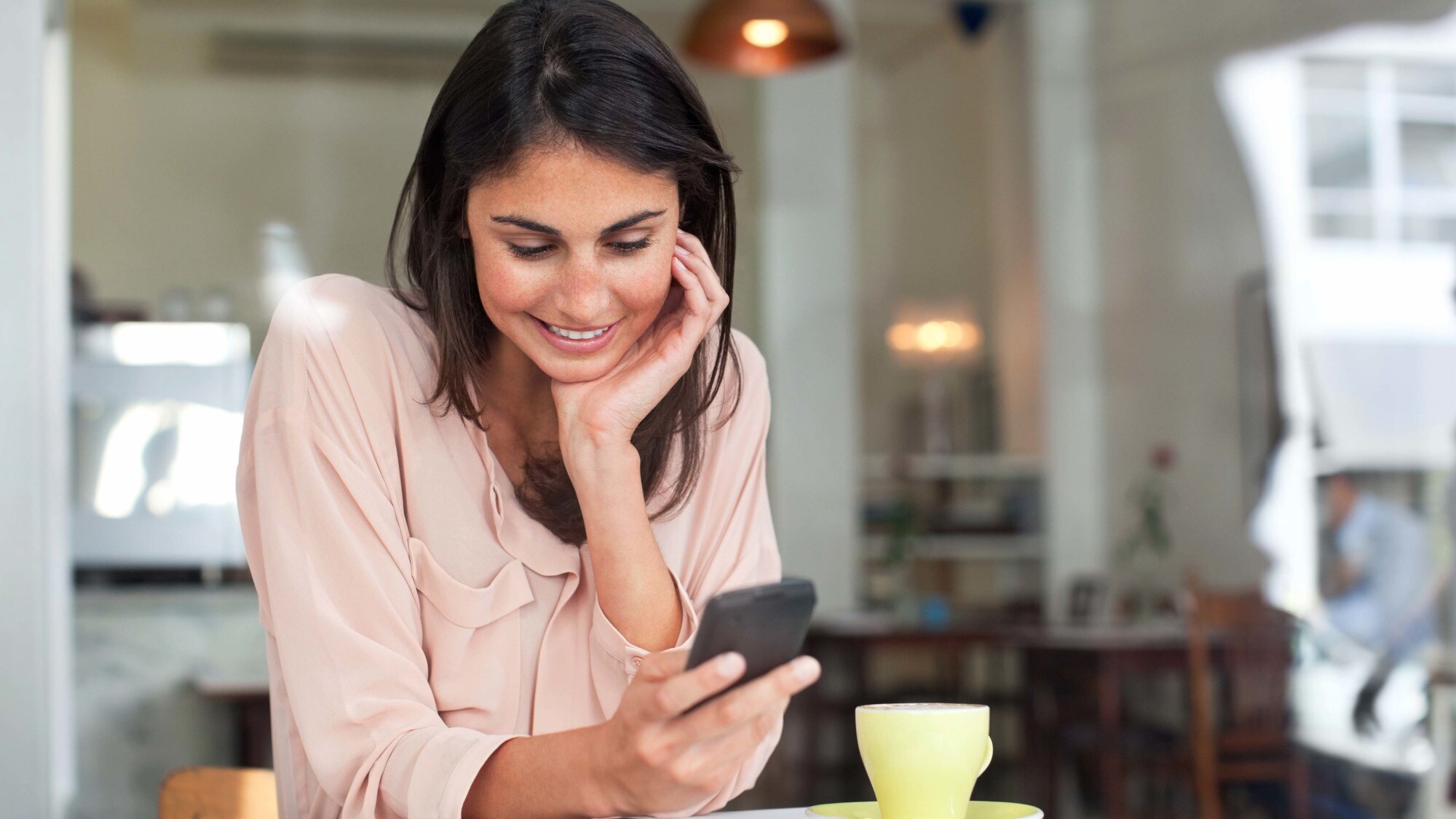 Eine junge Frau an einem Tisch in einem Café schaut lächelnd auf ihr Smartphone