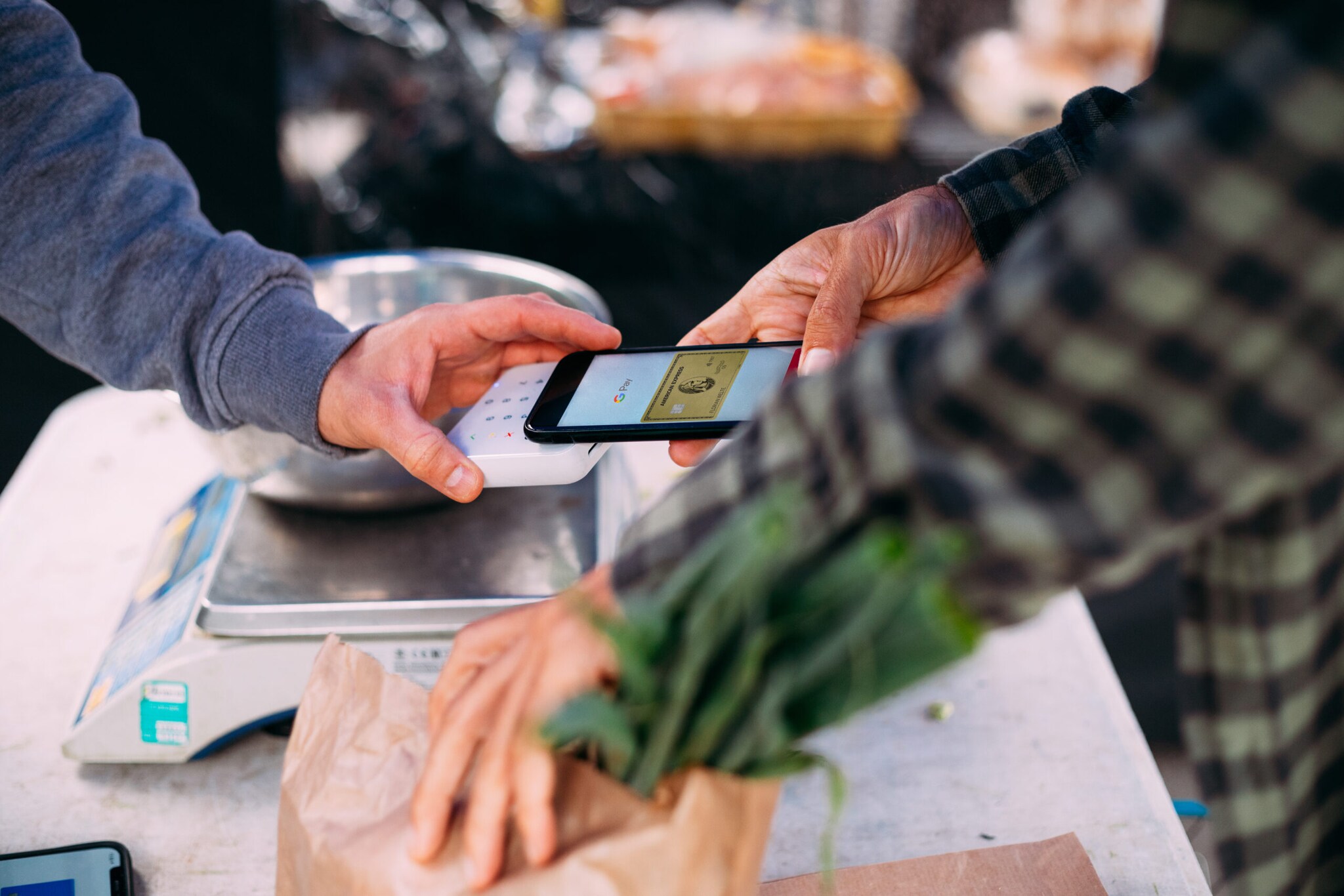 Kontaktlose Zahlung mit einer goldenen American Express Kreditkarte auf einem Smartphone, das an einem Marktstand an ein Kartenlesegerät gehalten wird