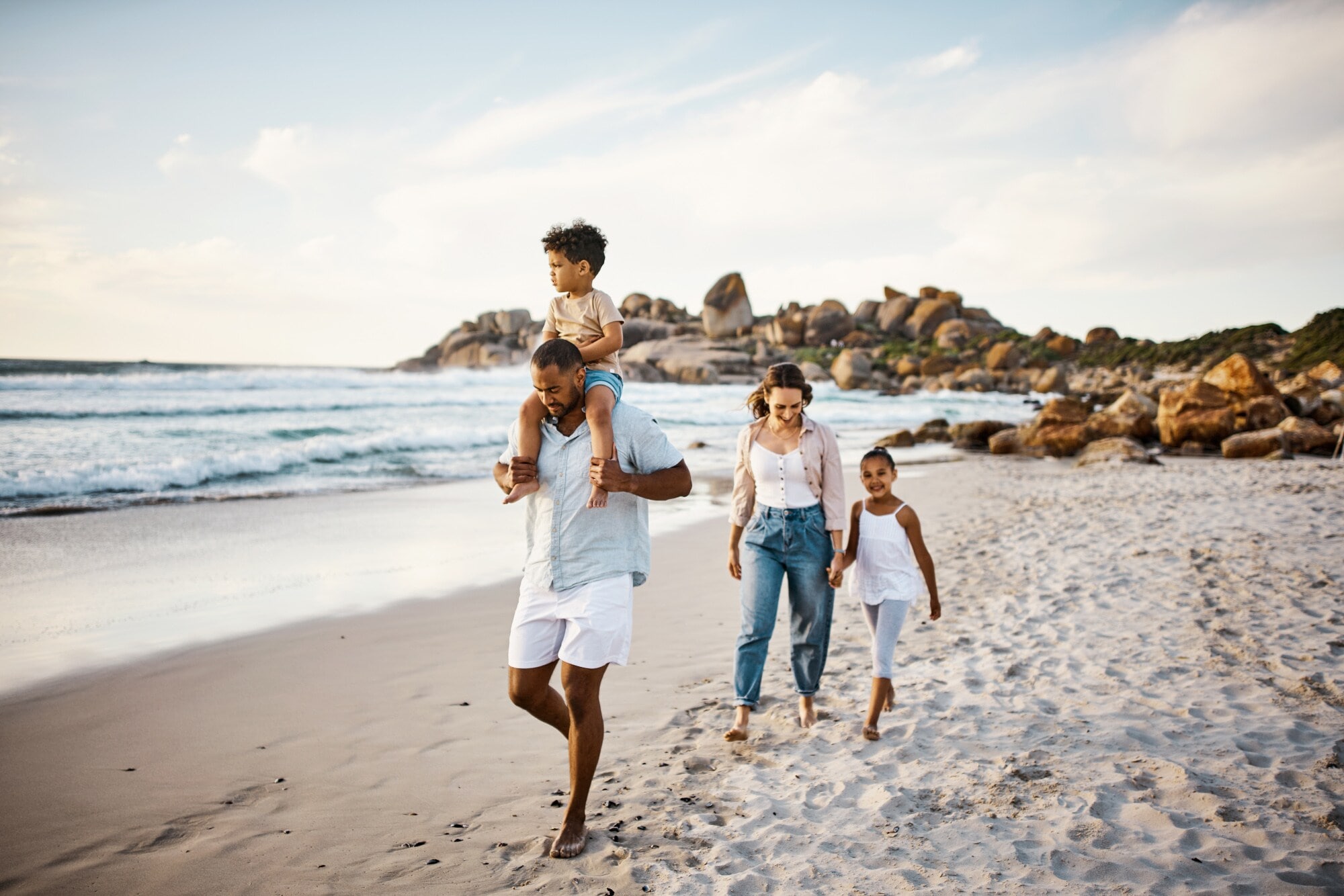 Eine junge Familie mit zwei Kindern spaziert auf einem Sandstrand am Meer entlang Eine junge Familie mit zwei Kindern spaziert auf einem Sandstrand am Meer entlang