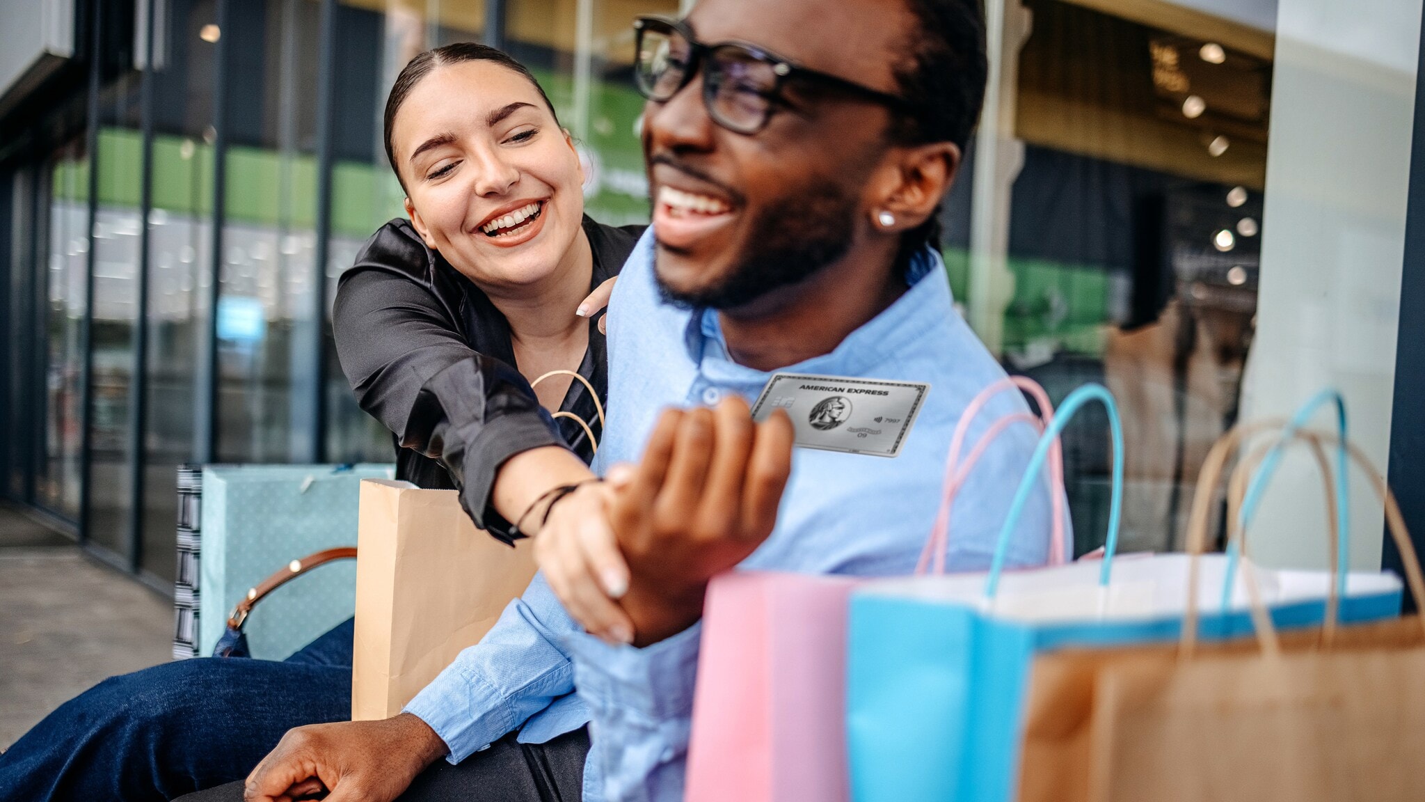 Eine Frau und ein Mann sitzen gut gelaunt neben Einkaufstüten auf einer Bank vor einem Geschäft, sie greift nach einer silbernen Kreditkarte von American Express in seiner Hand. Eine Frau und ein Mann sitzen gut gelaunt neben Einkaufstüten auf einer Bank vor einem Geschäft, sie greift nach einer silbernen Kreditkarte von American Express in seiner Hand.