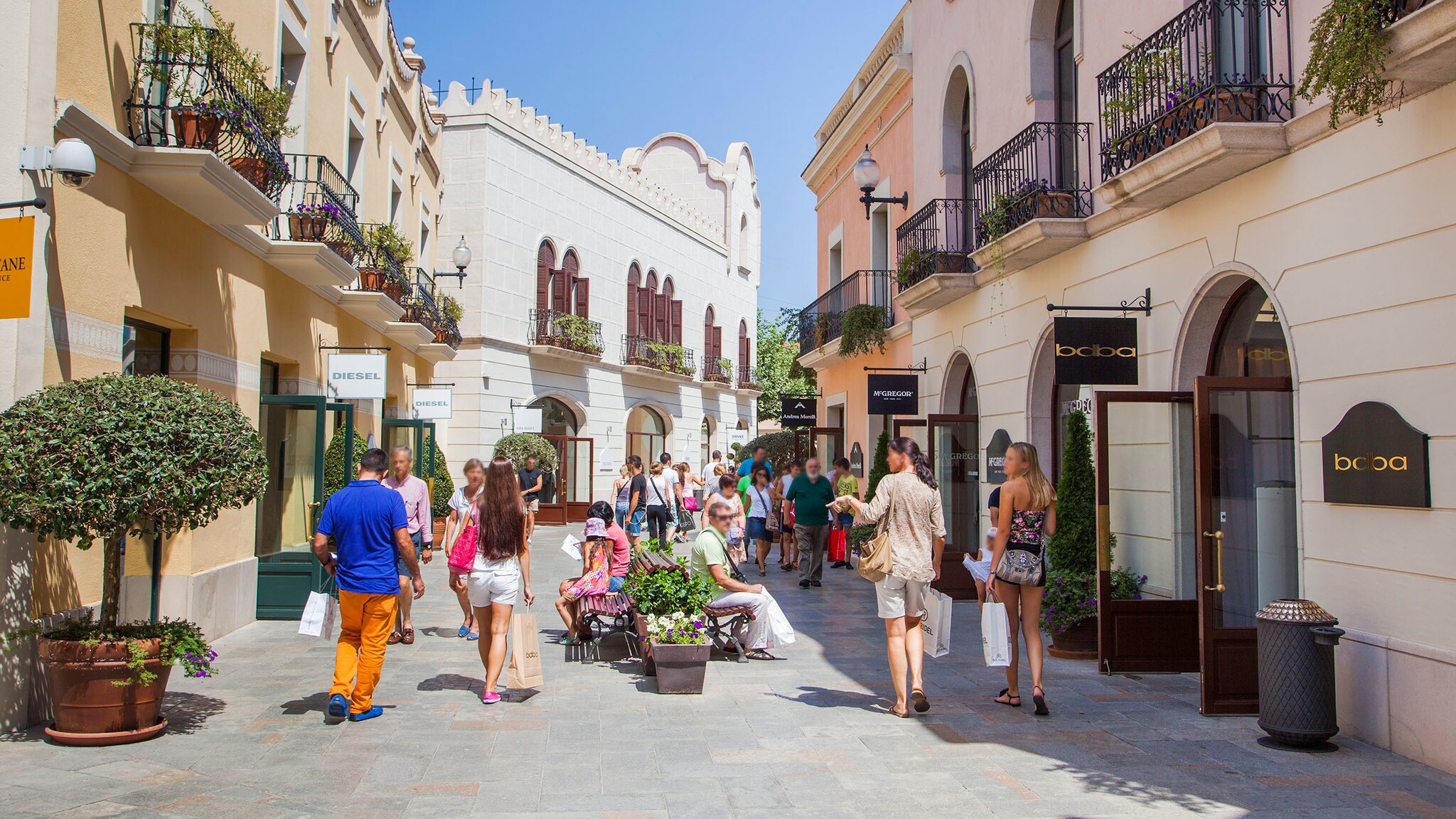Personen laufen durch eine Fußgängerzone mit Sitzbänken vorbei an Geschäften in einem Freiluft-Einkaufszentrum bei Sonnenschein. Personen laufen durch eine Fußgängerzone mit Sitzbänken vorbei an Geschäften in einem Freiluft-Einkaufszentrum bei Sonnenschein.