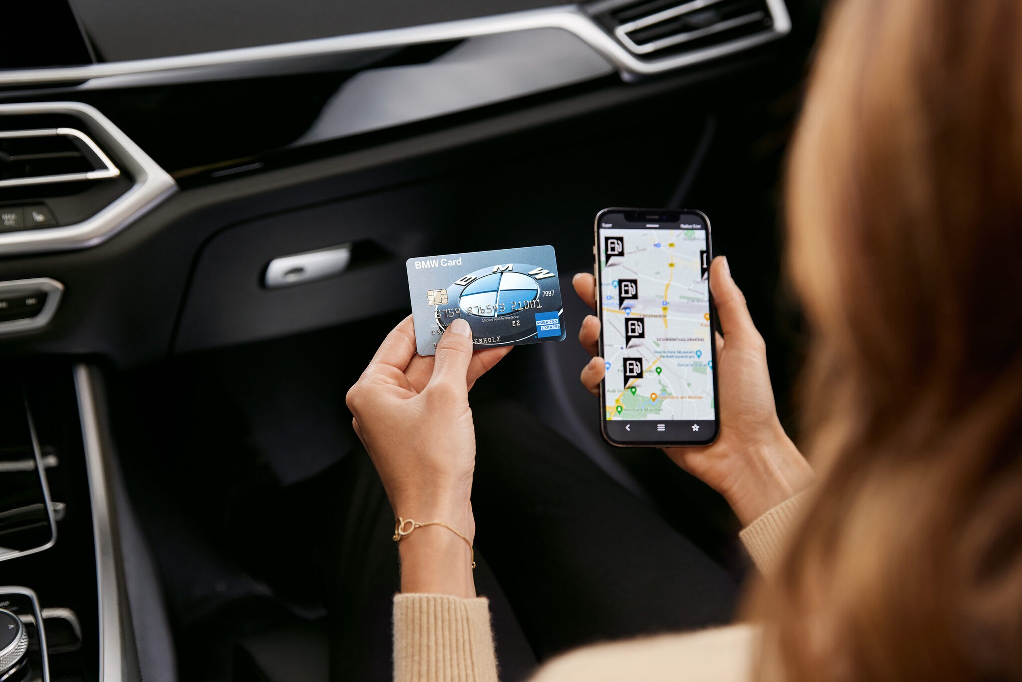 Eine Frau in einem Auto hält eine BMW Card von American Express neben ein Smartphone mit einer geöffneter App, die Tankstellen anzeigt