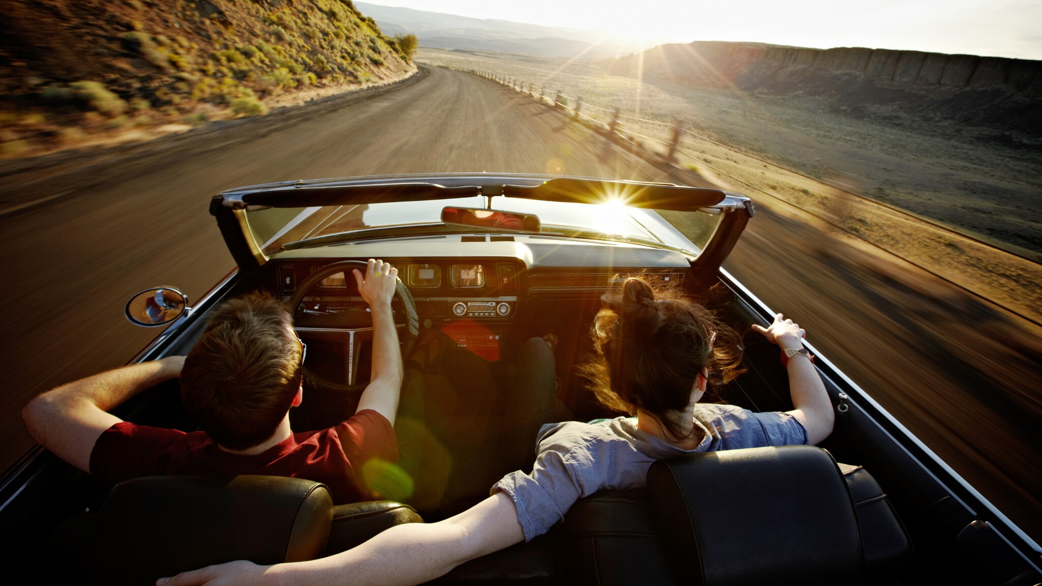 Rückansicht eines Paares in einem offenen Cabriolet während der Fahrt auf einer Straße durch eine Wüstenlandschaft bei Sonnenuntergang. Rückansicht eines Paares in einem offenen Cabriolet während der Fahrt auf einer Straße durch eine Wüstenlandschaft bei Sonnenuntergang.