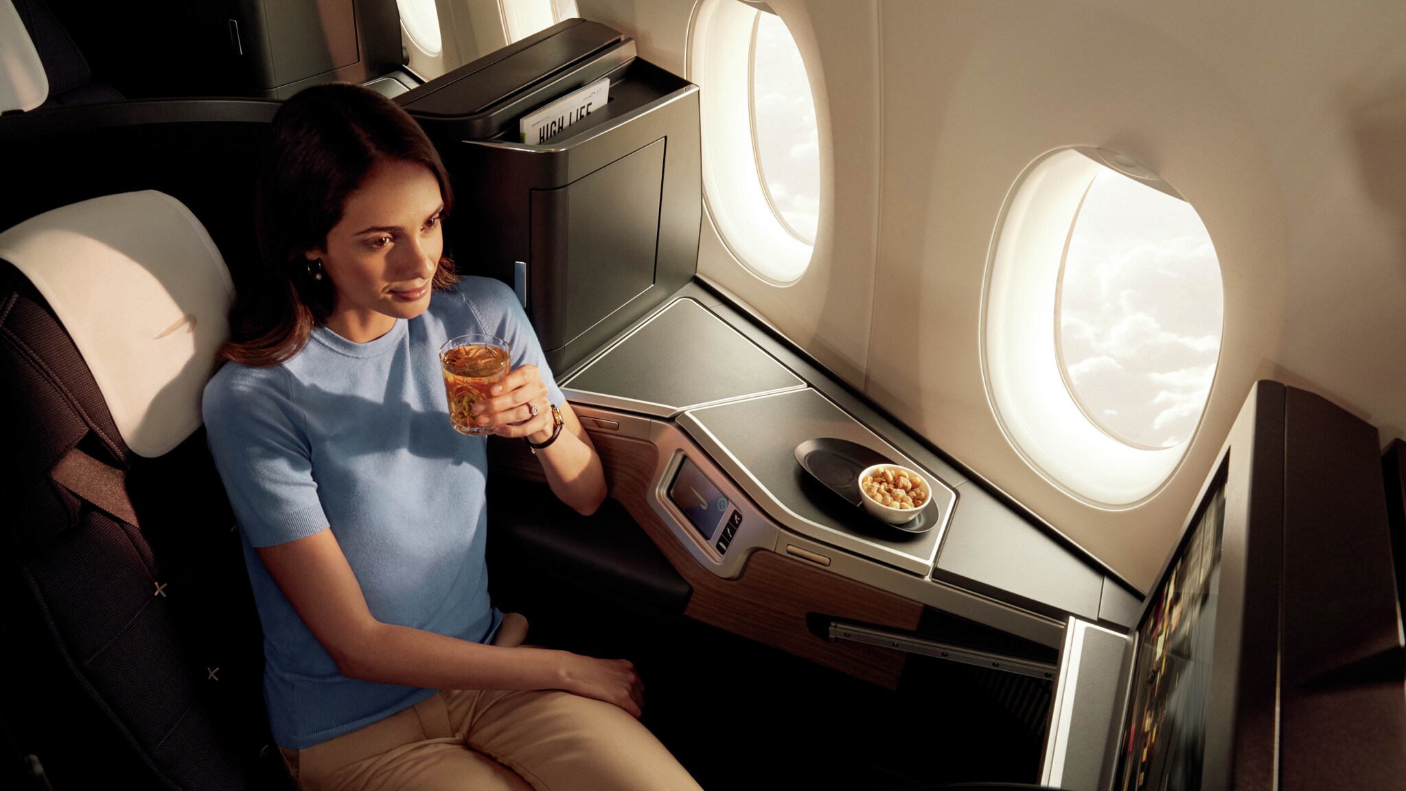 Eine Frau sitzt entspannt mit einem Getränk in der Business Class eines Flugzeugs.