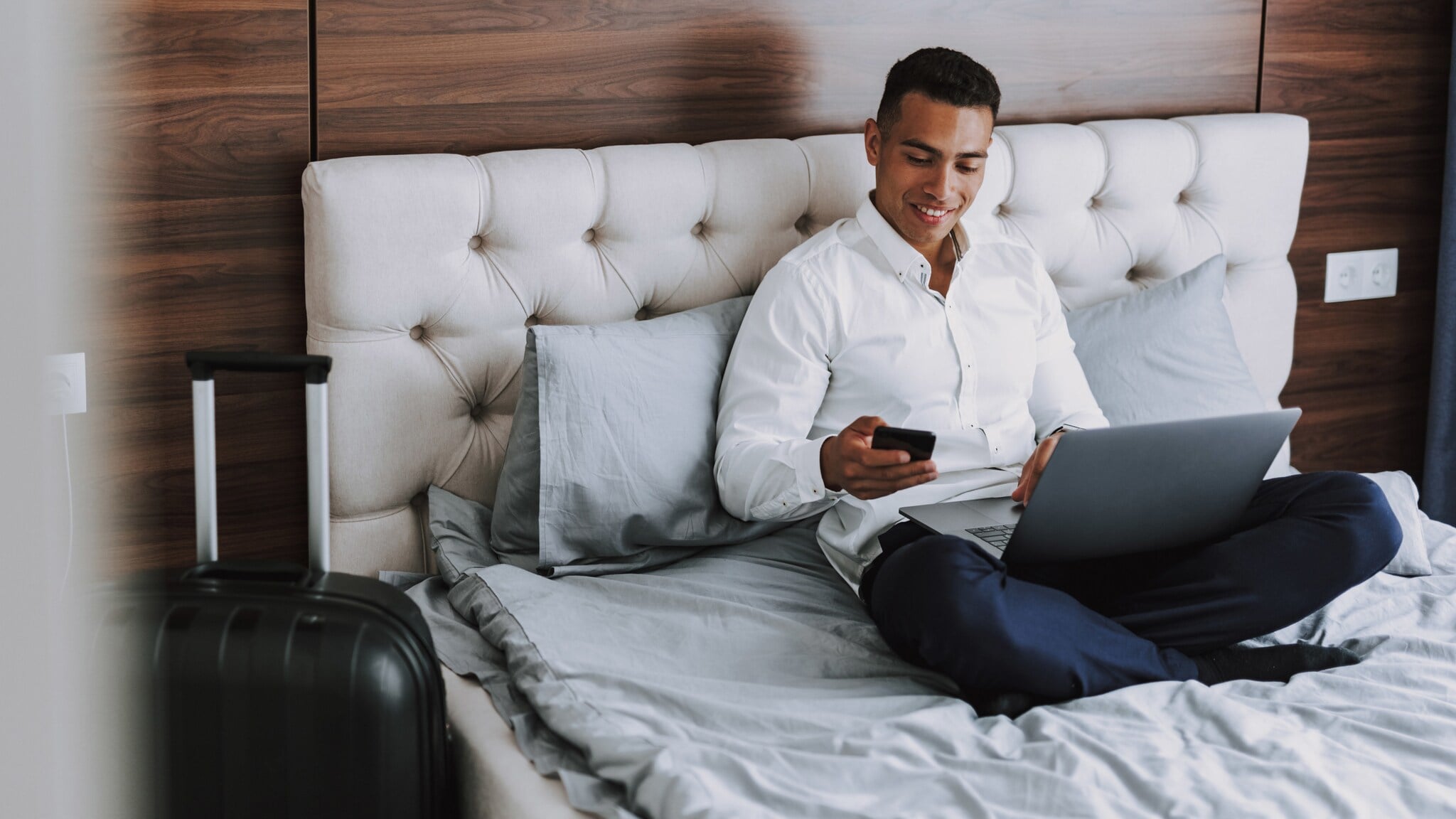 Mann sitzt in Hemd und Anzughose im Schneidersitz auf einem Hotelbett und telefoniert, auf dem Schoß einen Laptop. Mann sitzt in Hemd und Anzughose im Schneidersitz auf einem Hotelbett und telefoniert, auf dem Schoß einen Laptop.