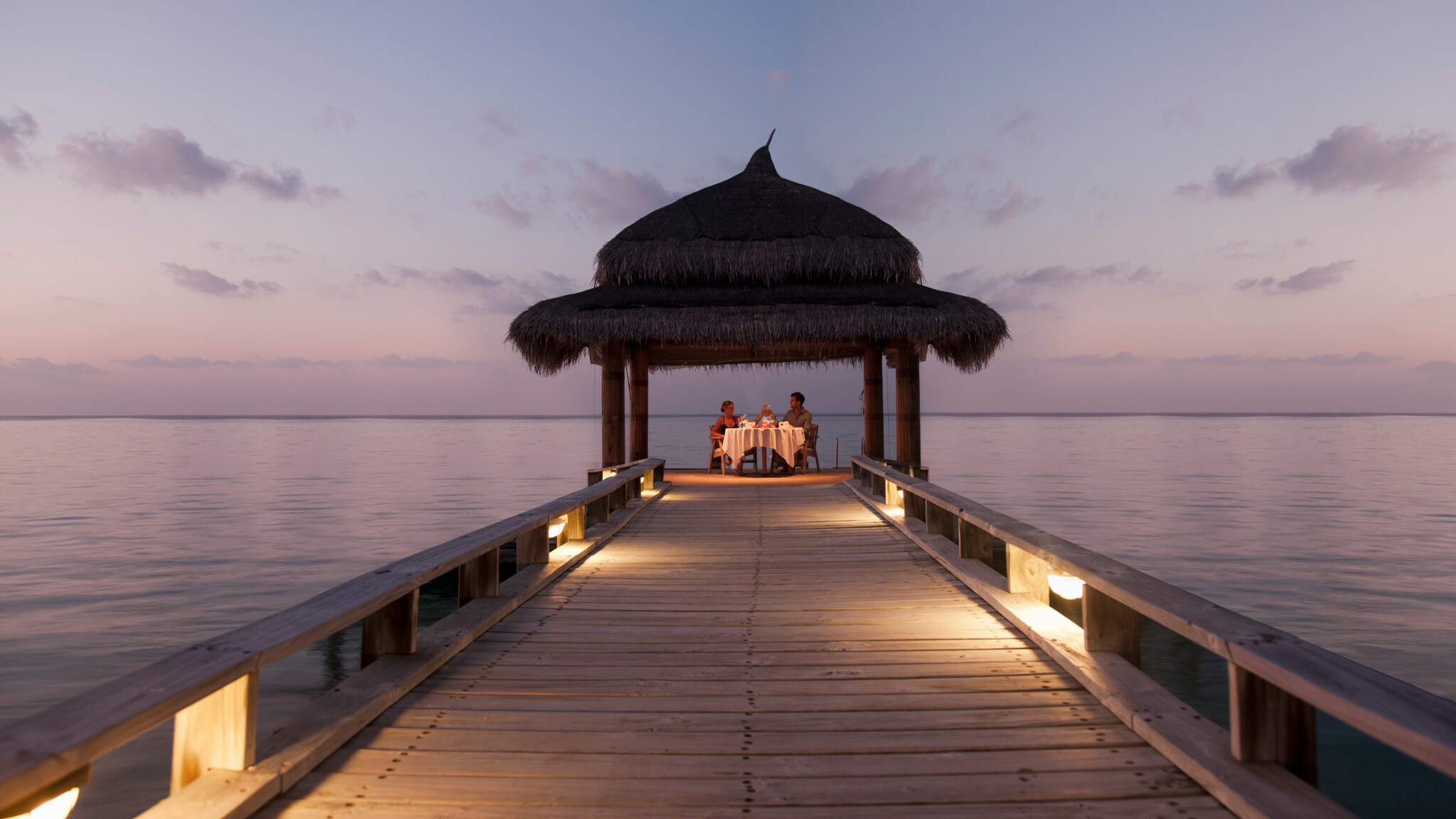 Ein Paar beim Candle-Light-Dinner unter einem Pavillon am Ende eines Stegs am Meer.