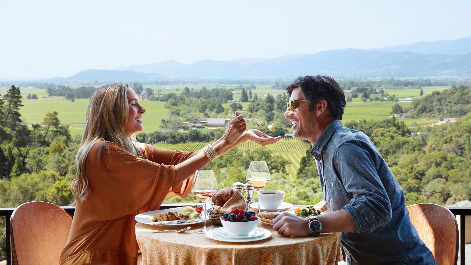 Ein Paar sitzt auf einer Terrasse beim Frühstück vor grünem Landschaftspanorama, sie reicht ihm einen Probierbissen auf der Gabel über den Tisch.