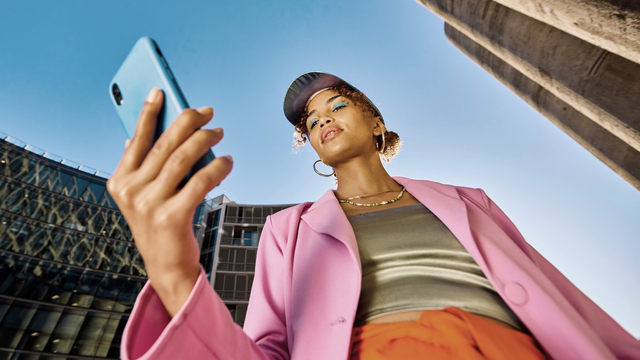 Eine stylishe junge Frau schaut auf ihr Smartphone in urbaner Umgebung unter blauem Himmel.