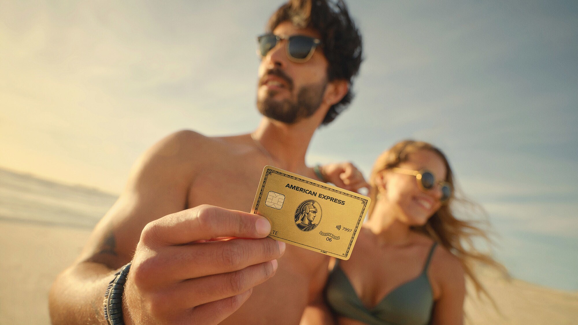 Ein Paar am Strand in Unschärfe, er hält eine goldene Kreditkarte von American Express aus Metall hoch.