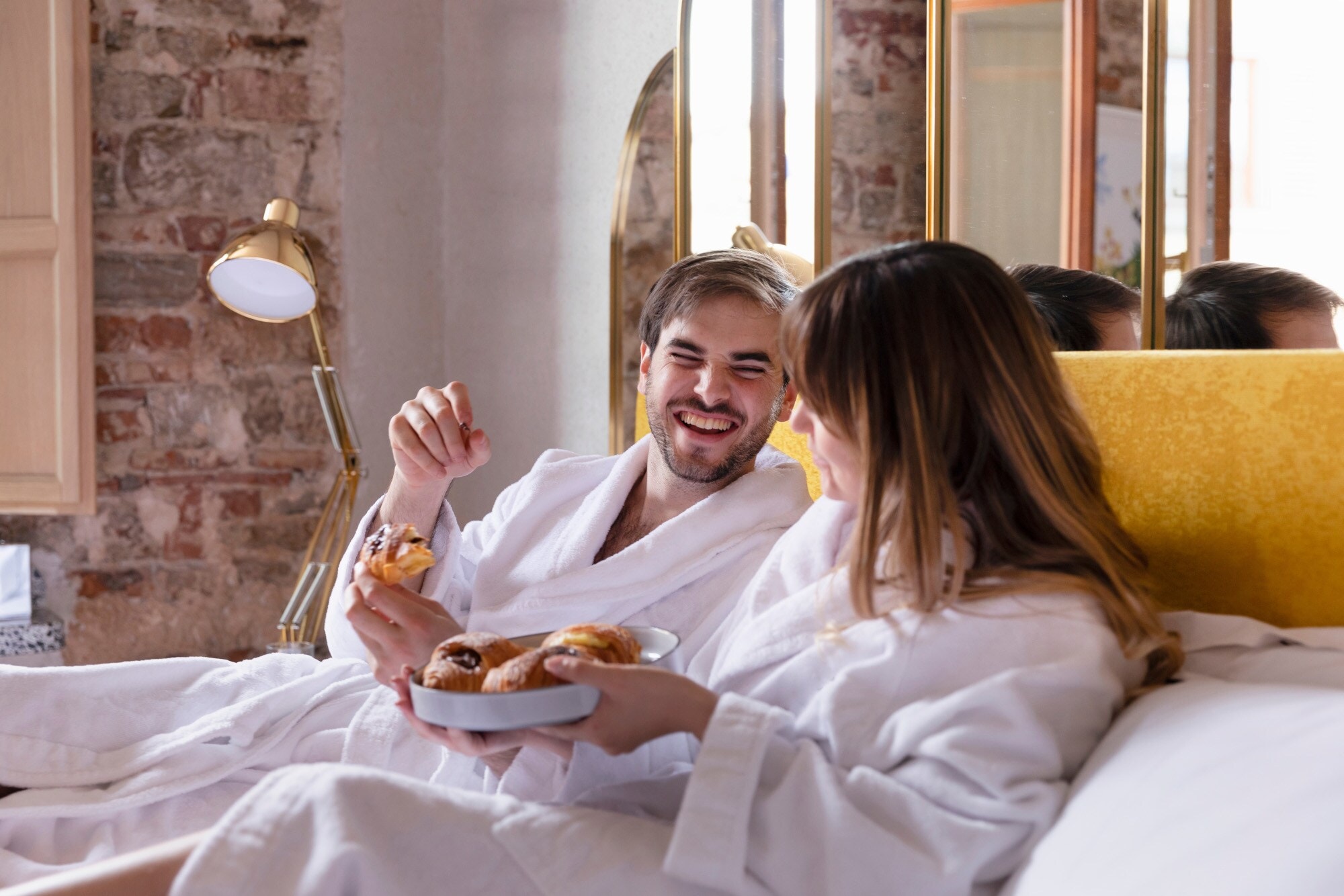 Ein gut gelauntes Paar in weißen Bademänteln mit Croissants in den Händen liegt auf einem Bett. Ein gut gelauntes Paar in weißen Bademänteln mit Croissants in den Händen liegt auf einem Bett.