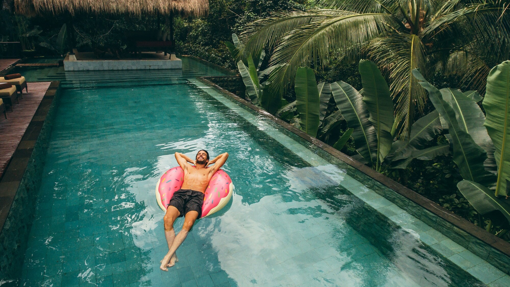 Ein Mann schwimmt entspannt rücklings auf einer pinken Luftmatratze in einem Hotelpool in tropischer Umgebung. Ein Mann schwimmt entspannt rücklings auf einer pinken Luftmatratze in einem Hotelpool in tropischer Umgebung.