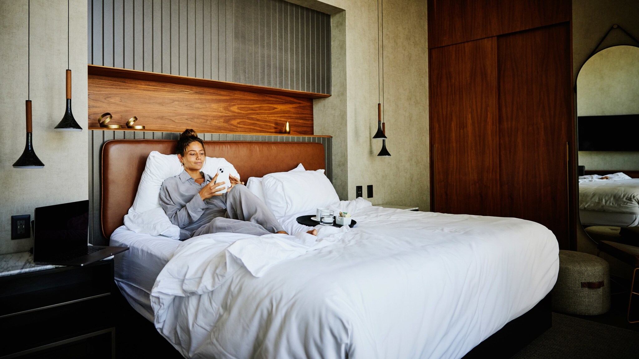 Eine junge Frau im Pyjama liegt auf einem großen Bett und schaut auf ihr Smartphone in einem stilvollen Hotelzimmer bei Tageslicht. Eine junge Frau im Pyjama liegt auf einem großen Bett und schaut auf ihr Smartphone in einem stilvollen Hotelzimmer bei Tageslicht.