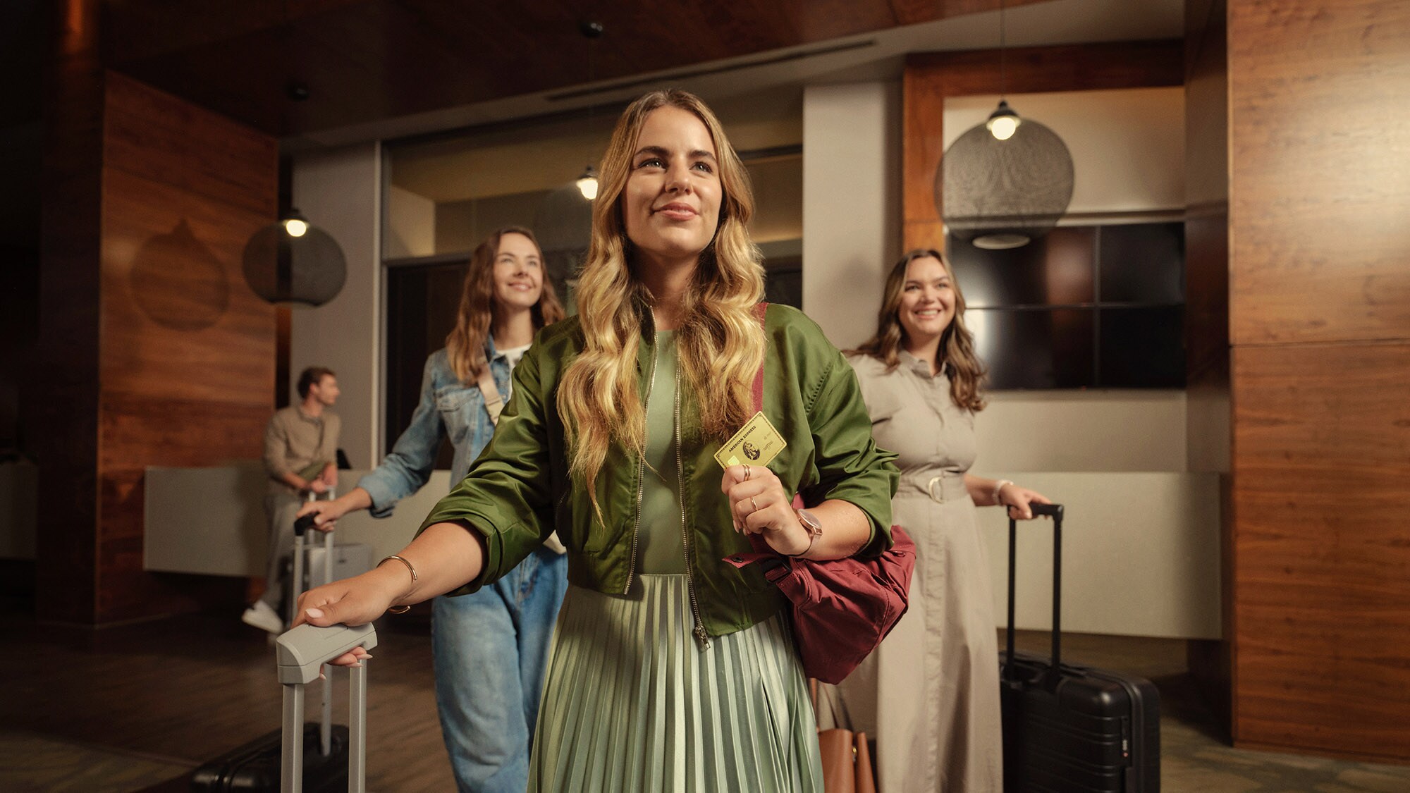Drei junge Frauen gehen fröhlich mit Trolleys durch eine Hotellobby, die Frau vorne hält eine goldene Kreditkarte von American Express in ihrer Hand. Drei junge Frauen gehen fröhlich mit Trolleys durch eine Hotellobby, die Frau vorne hält eine goldene Kreditkarte von American Express in ihrer Hand.