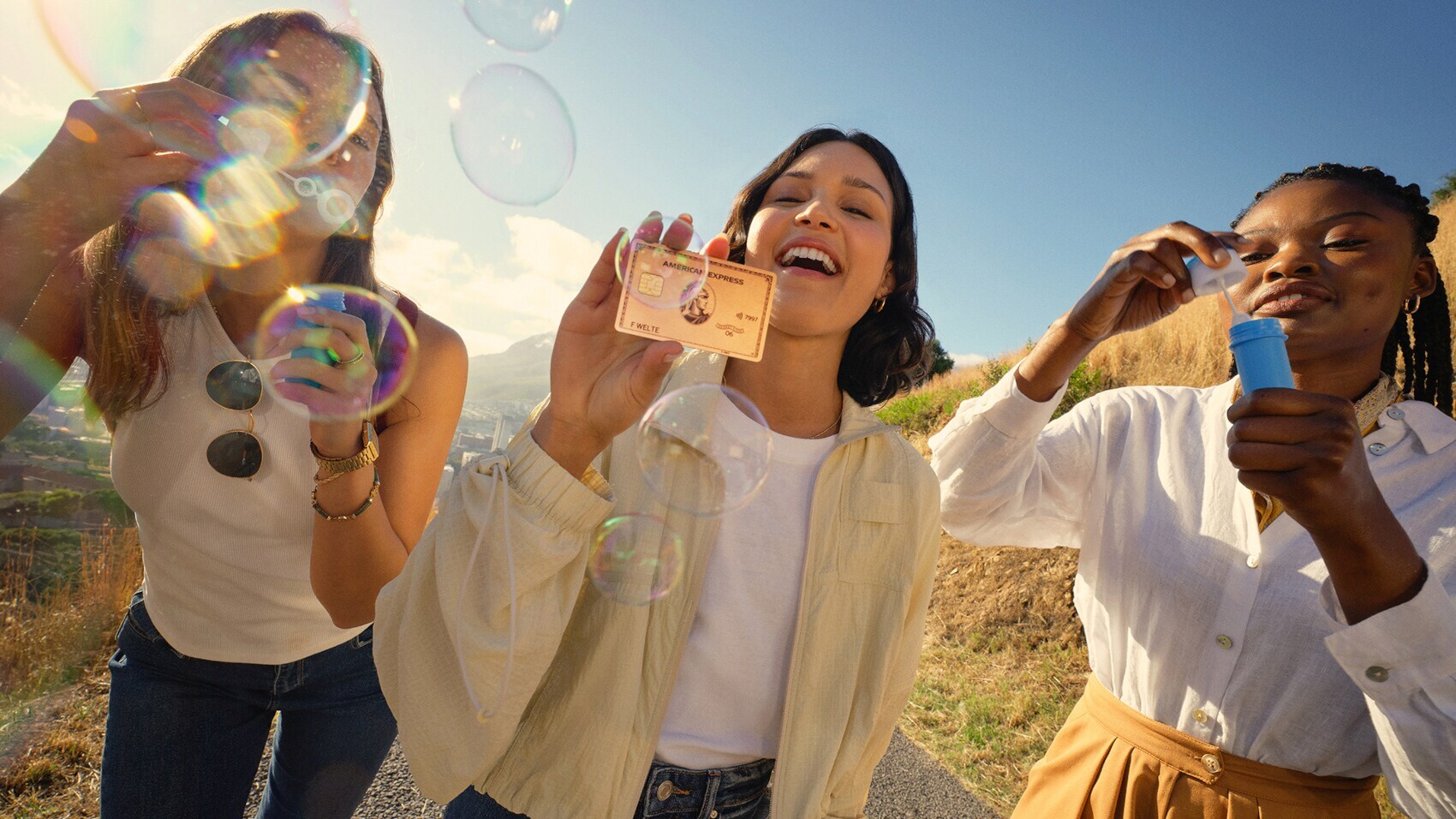 Drei Frauen unter freiem Himmel bei Sonnenschein, von denen zwei Seifenblasen blasen und eine die roséfarbene Kreditkarte von American Express präsentiert.