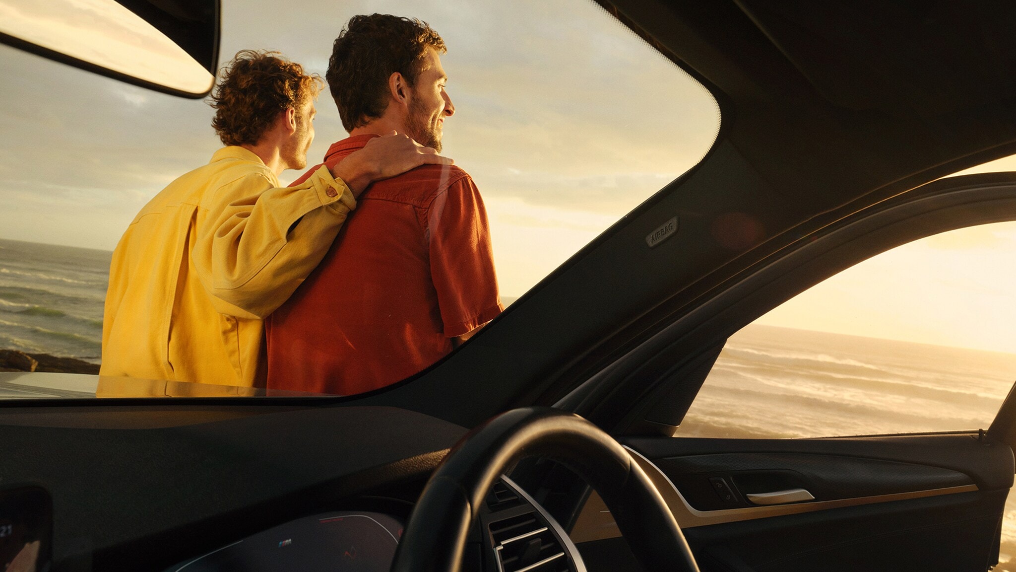 Rückansicht von zwei Männern, die an ein Auto gelehnt sind. Man sieht von dem Auto einen Teil des Innenraums, die Männer blicken auf das Meer hinaus und lächeln. Rückansicht von zwei Männern, die an ein Auto gelehnt sind. Man sieht von dem Auto einen Teil des Innenraums, die Männer blicken auf das Meer hinaus und lächeln.
