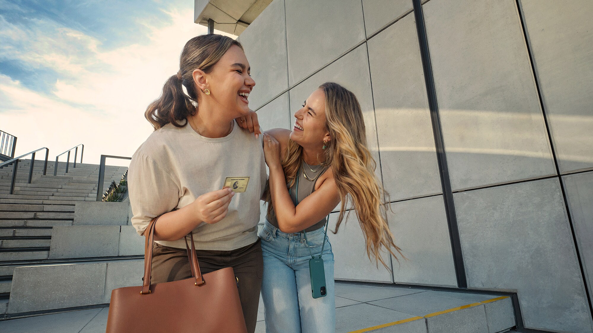 Zwei sich umarmende, lachende junge Frauen, eine hält eine goldene Kreditkarte von American Express in der Hand.