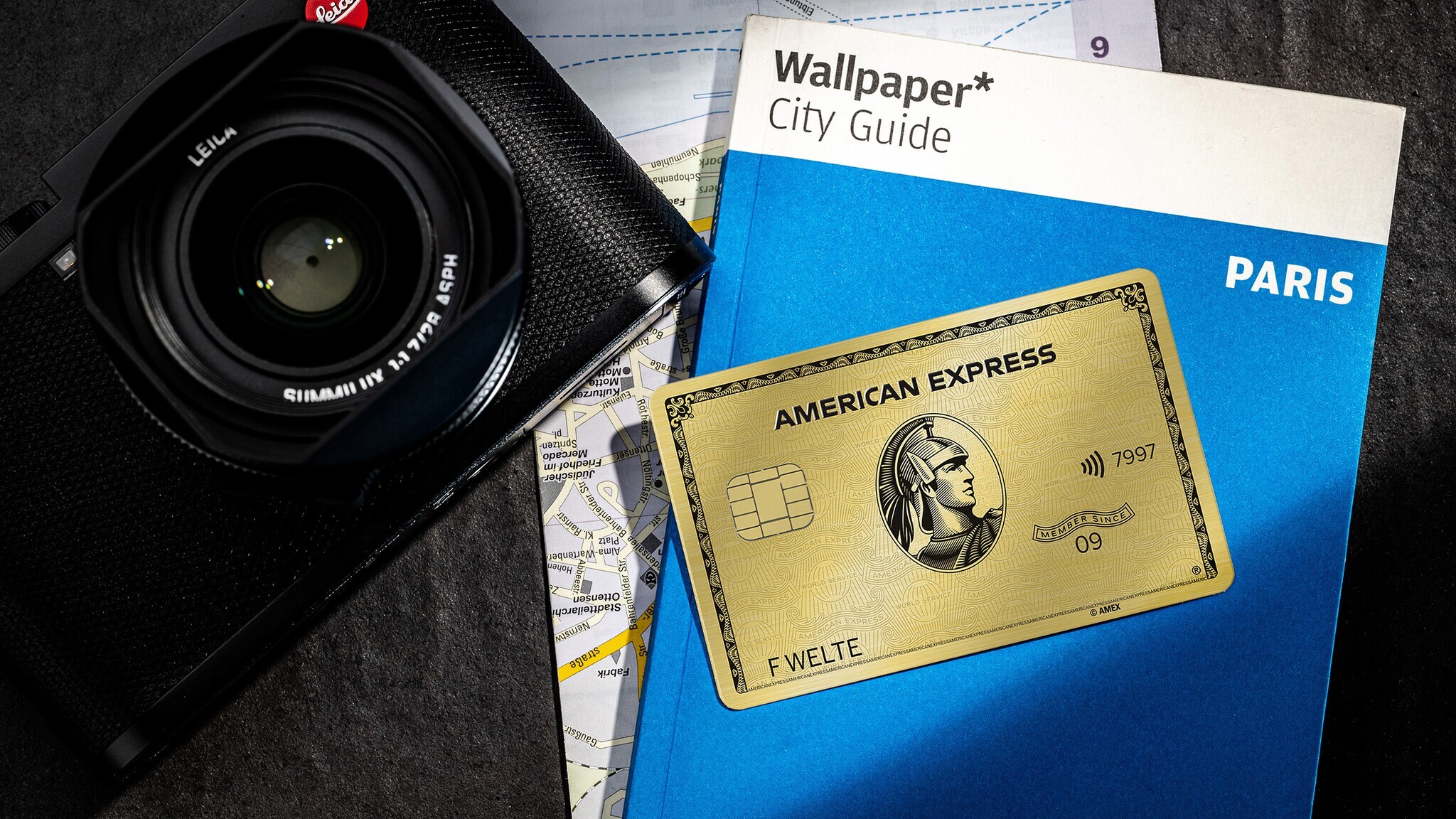 Aufsicht einer goldenen Kreditkarte von American Express auf einem Reiseführer mit Stadtplan neben einer Kamera.