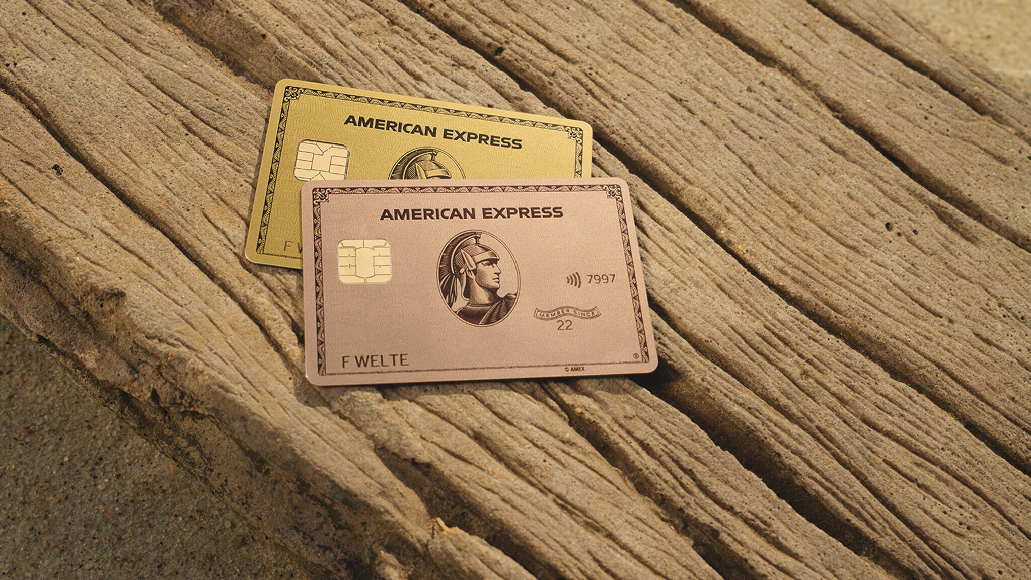 Eine American Express Gold Card und eine Gold Card Rosé liegen auf einem Stück Treibholz bei Sonnenuntergang. Eine American Express Gold Card und eine Gold Card Rosé liegen auf einem Stück Treibholz bei Sonnenuntergang.