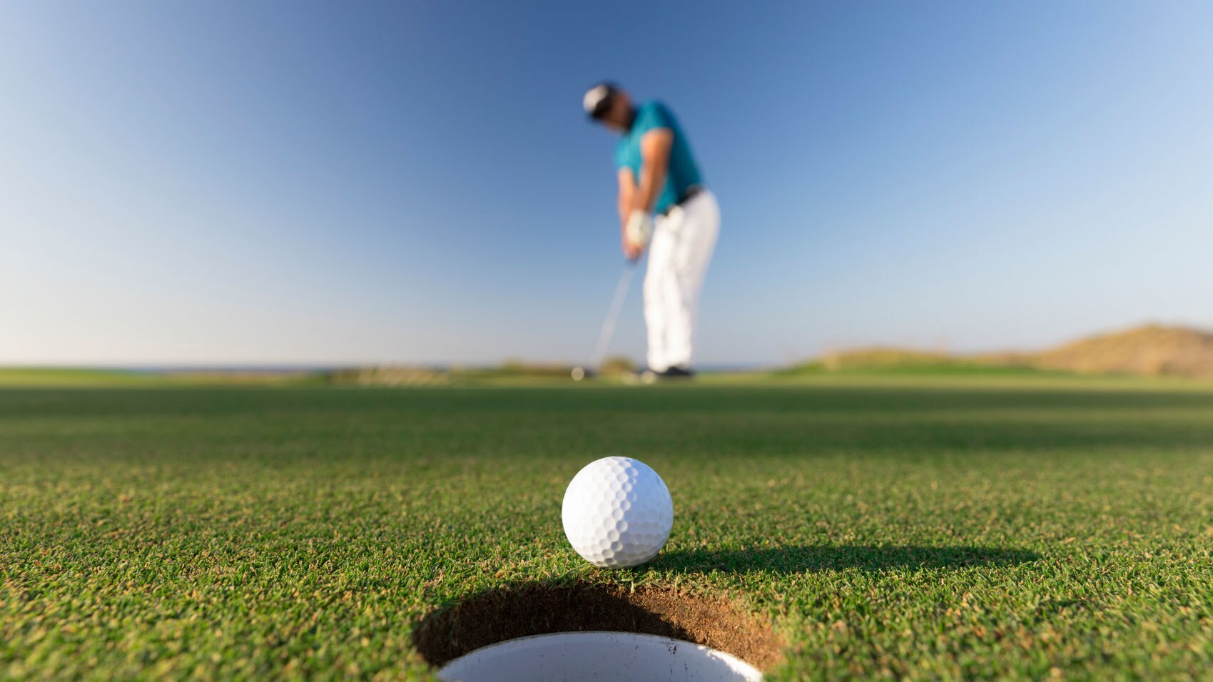 Nahaufnahme eines Golfballs an einem Loch auf einer Rasenfläche, im Hintergrund ein Mann mit Golfschläger in der Unschärfe vor blauem Himmel.