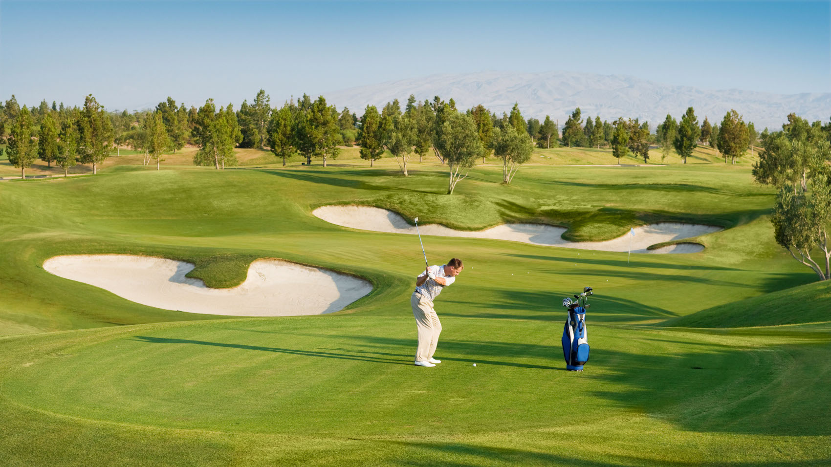 Ein Mann beim Abschlag auf einem hügeligen Golfplatz mit Sandbunkern bei Sonnenschein. Ein Mann beim Abschlag auf einem hügeligen Golfplatz mit Sandbunkern bei Sonnenschein.