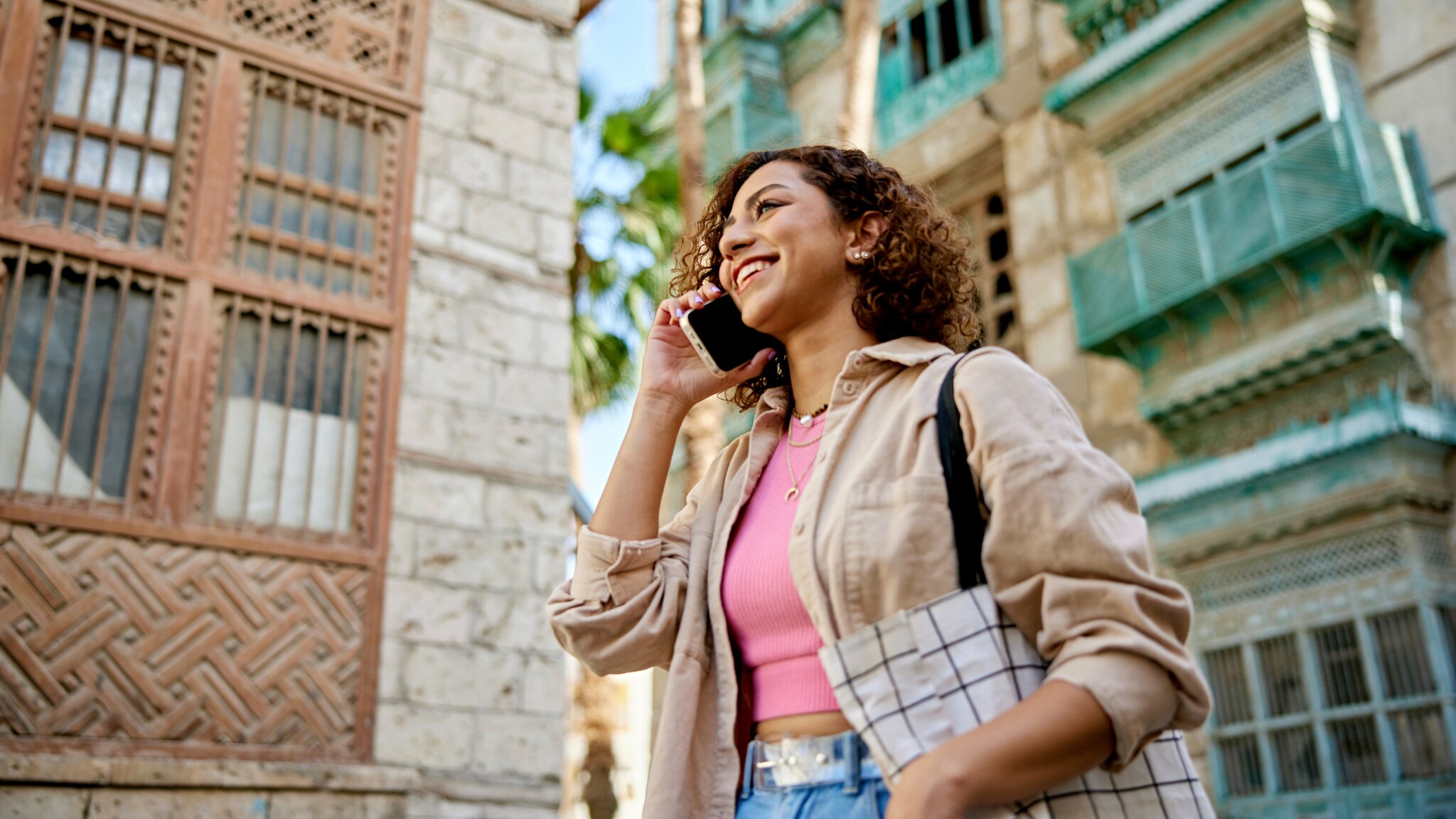 Eine lächelnde Frau telefoniert mit Smartphone auf einer Straße vor einer Häuserfront mit Palme.