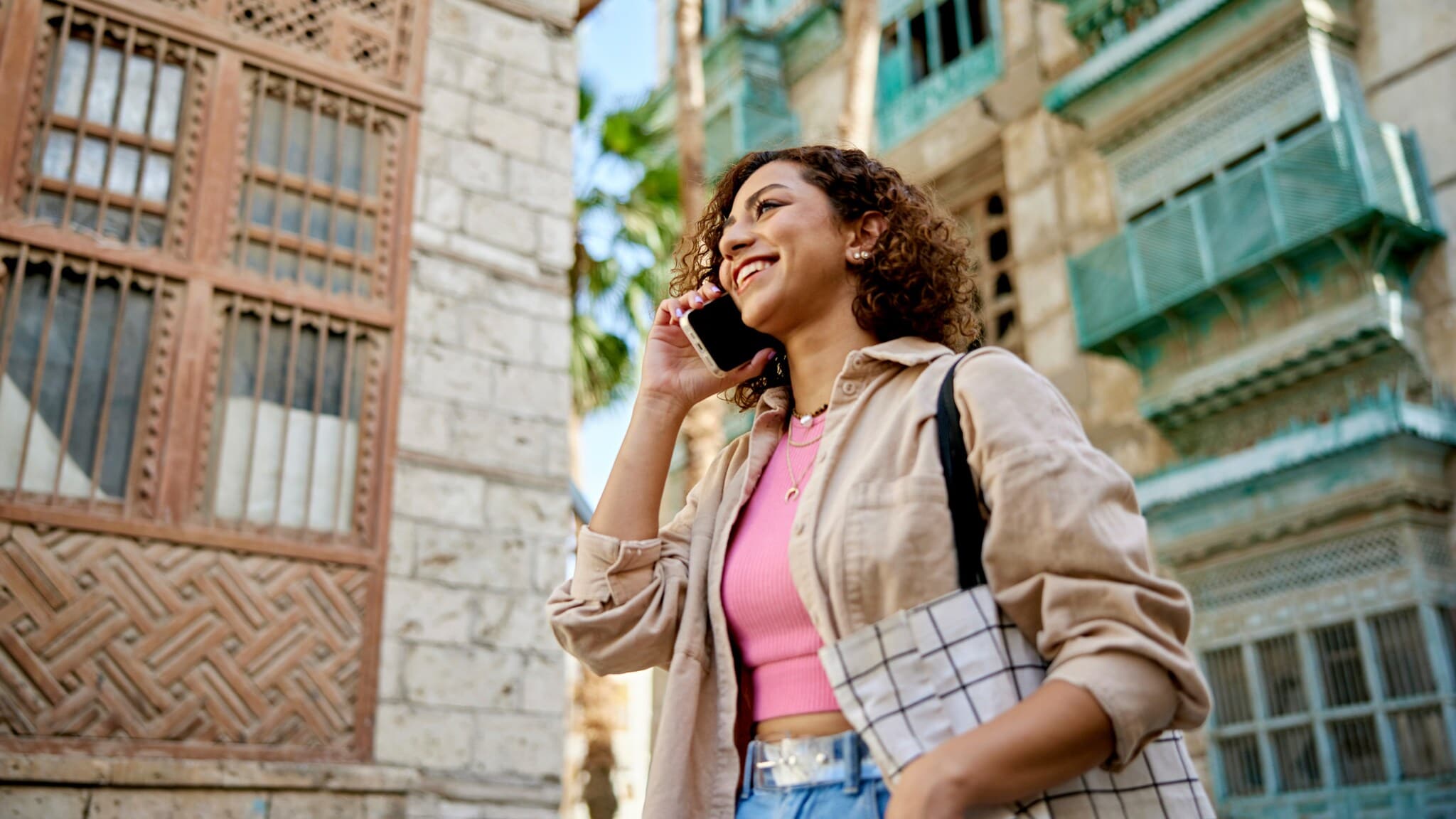 Eine lächelnde Frau telefoniert mit Smartphone auf einer Straße vor einer Häuserfront mit Palme. Eine lächelnde Frau telefoniert mit Smartphone auf einer Straße vor einer Häuserfront mit Palme.
