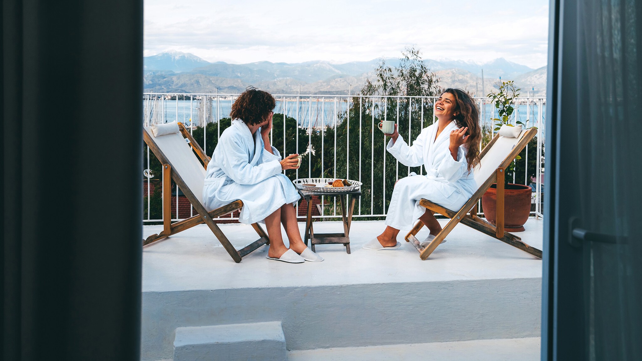 Zwei Freundinnen in weißen Bademänteln unterhalten sich bei Kaffeetrinken auf einem Balkon mit Blick auf einen See.