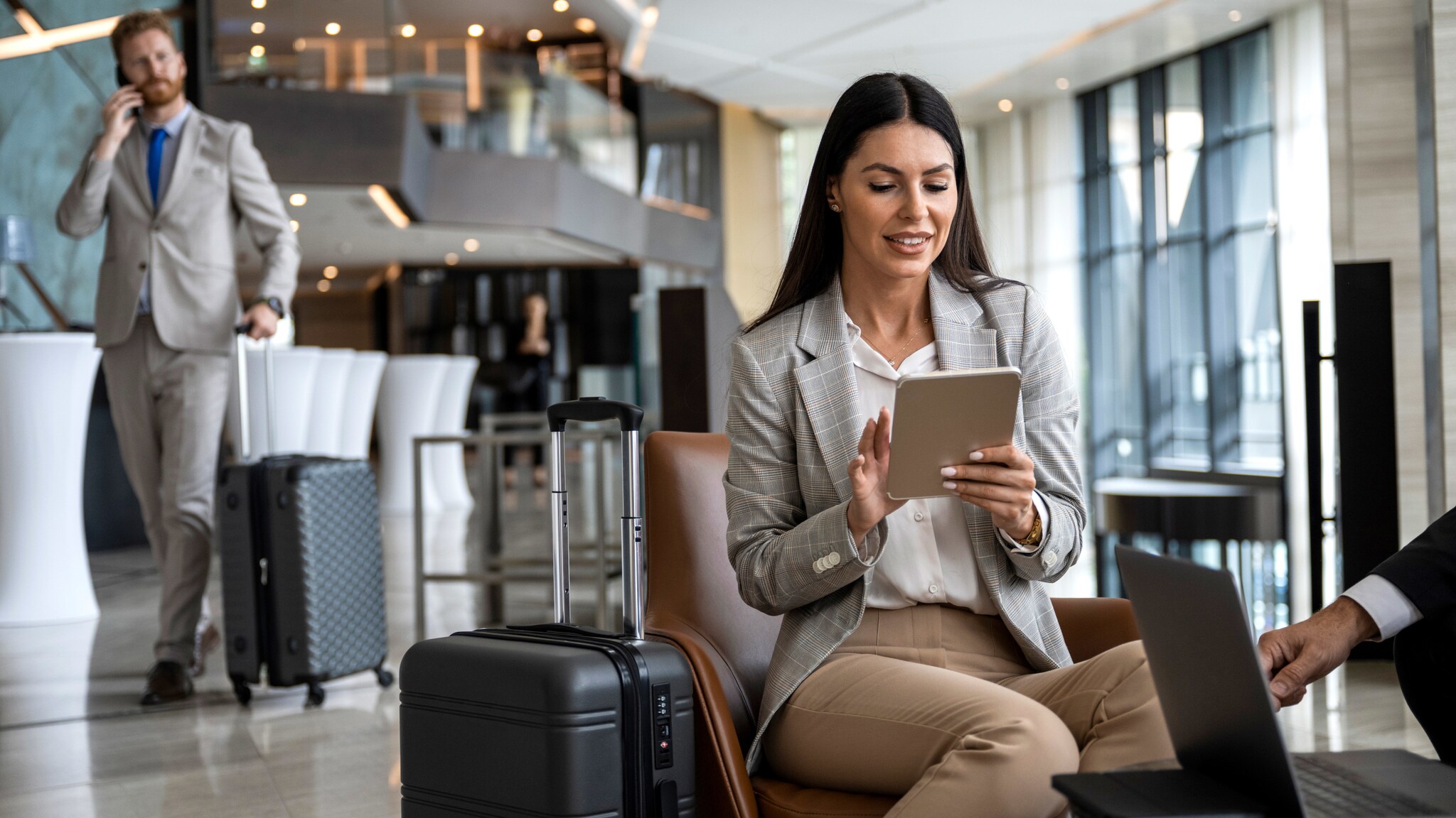 Eine Geschäftsfrau mit Koffer sitzt in einem Sessel und schaut auf ein Tablet in einer hellen Lounge, im Hintergrund läuft ein telefonierender Geschäftsmann mit Trolley. Eine Geschäftsfrau mit Koffer sitzt in einem Sessel und schaut auf ein Tablet in einer hellen Lounge, im Hintergrund läuft ein telefonierender Geschäftsmann mit Trolley.