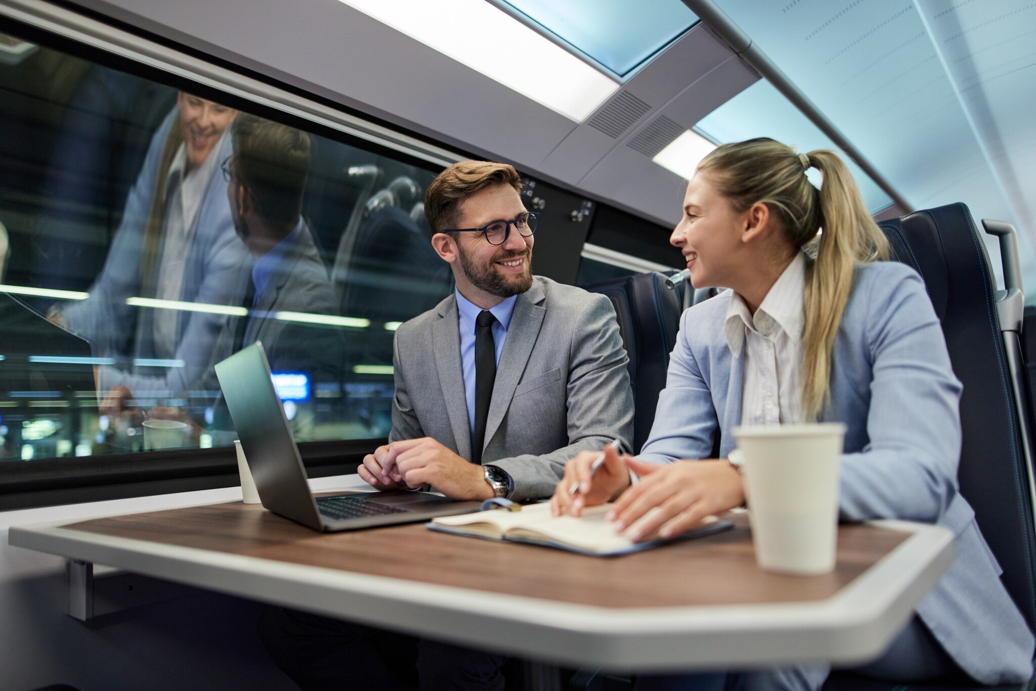 Ein Mann und eine Frau in Anzügen sitzen an einem Tisch in einem Zugabteil und unterhalten sich gut gelaunt vor einem Laptop.