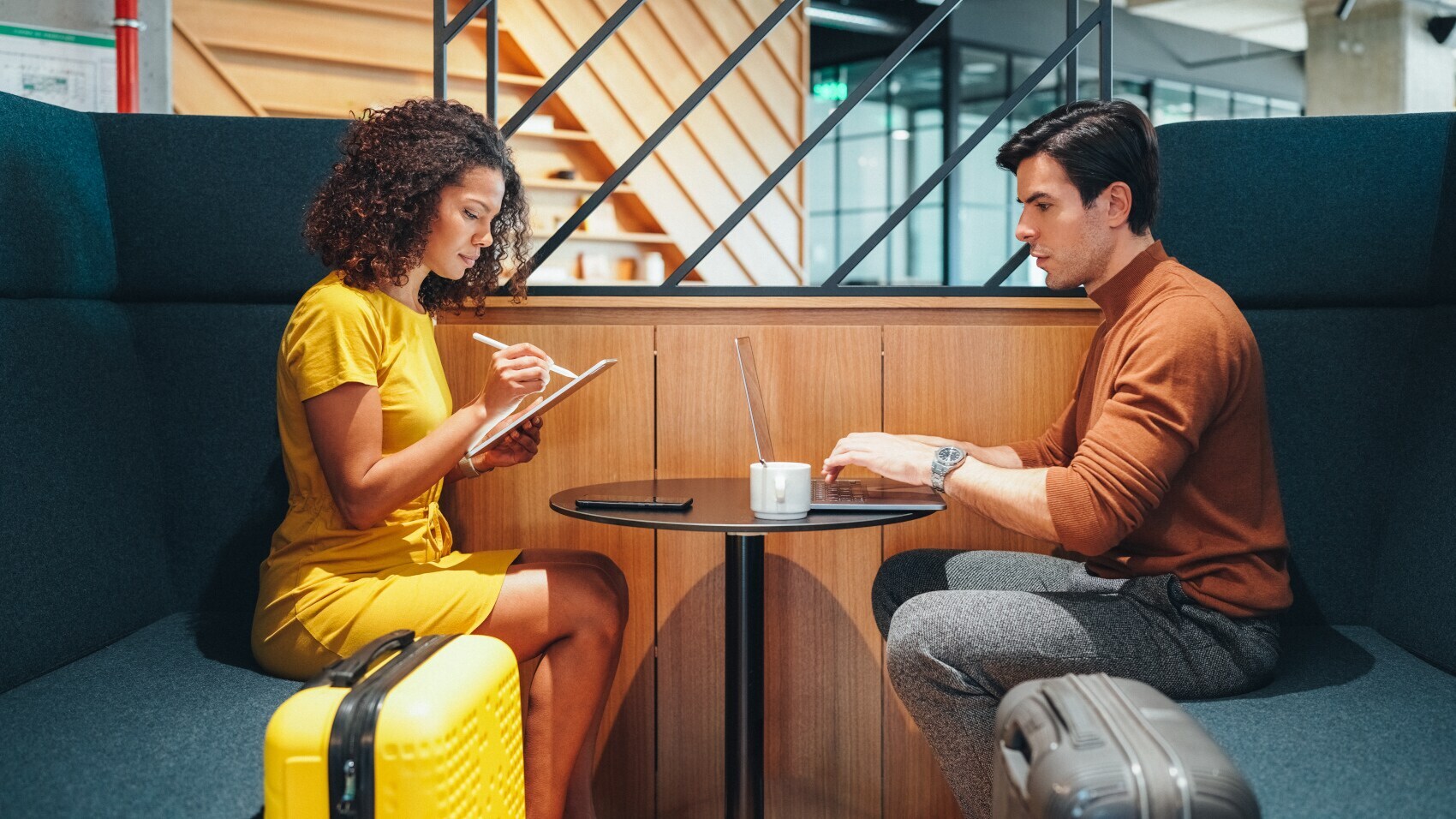 Ein Mann und eine Frau mit Koffern arbeiten an mobilen Geräten an einem Tisch in einer Flughafenlounge