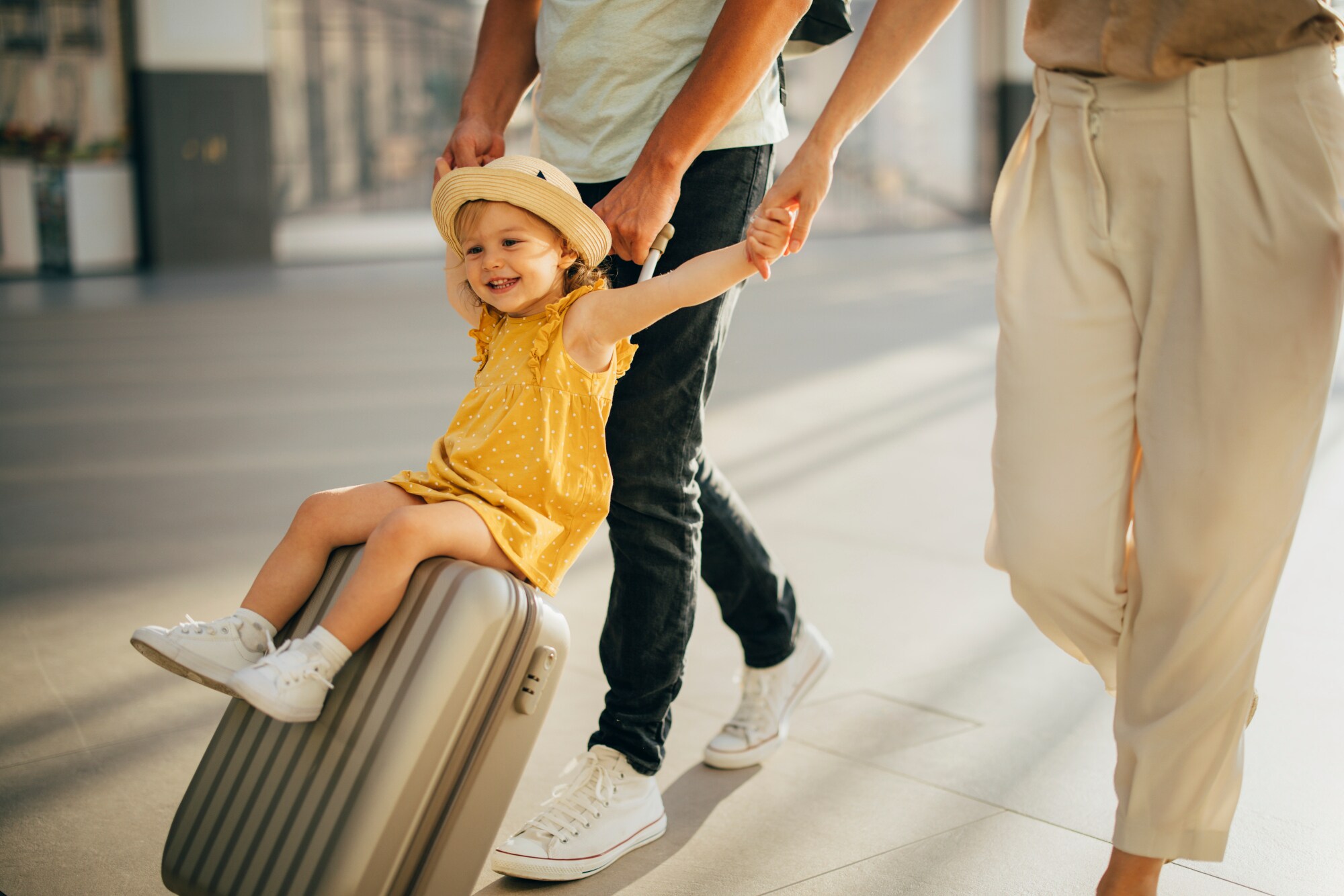 Ein kleines, fröhliches Mädchen im gelben Kleid sitzt an den Händen zweier Personen auf einem grauen Hartschalenkoffer.