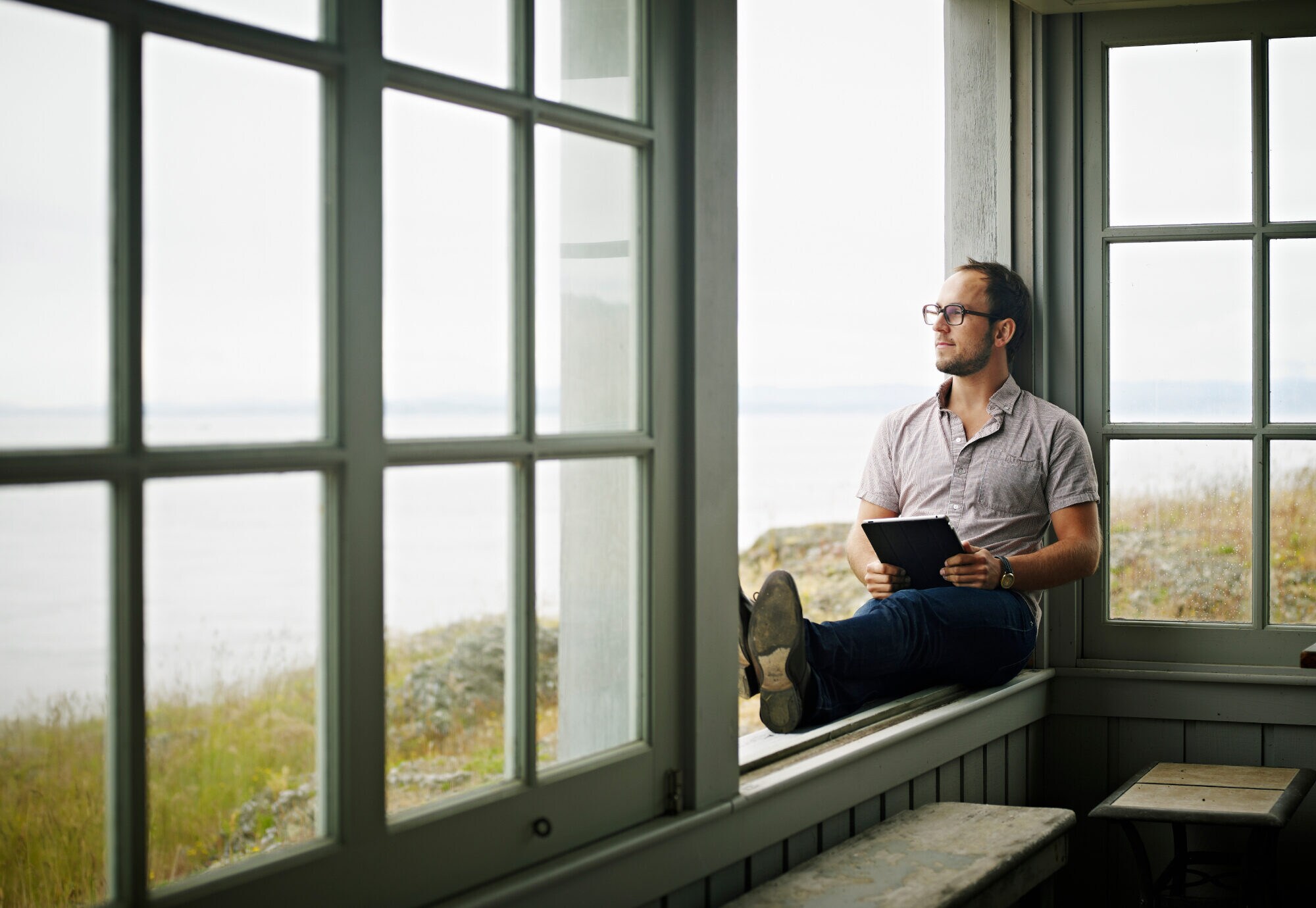 Ein Mann sitzt mit einem Tablet auf einer Fensterbank und blickt aufs Meer hinaus