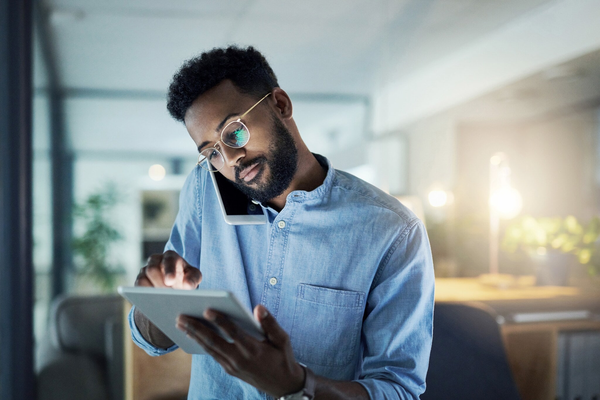 Ein junger Mann im Büro bedient ein Tablet in der Hand, während er ein Smartphone am Ohr eingeklemmt hat Ein junger Mann im Büro bedient ein Tablet in der Hand, während er ein Smartphone am Ohr eingeklemmt hat