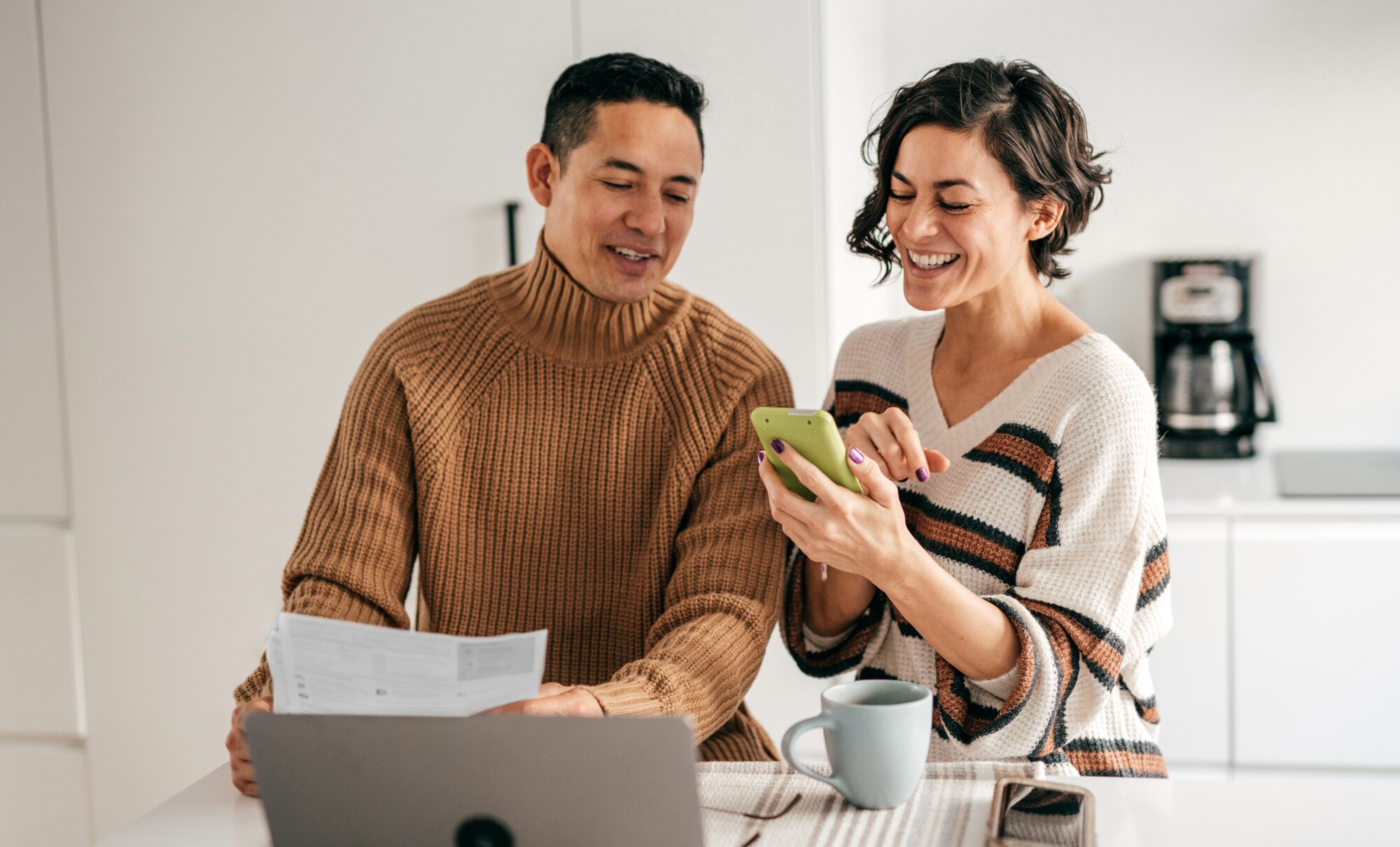 Ein Paar steht mit einer Abrechnung in der Küche und schaut gut gelaunt auf ein Smartphone