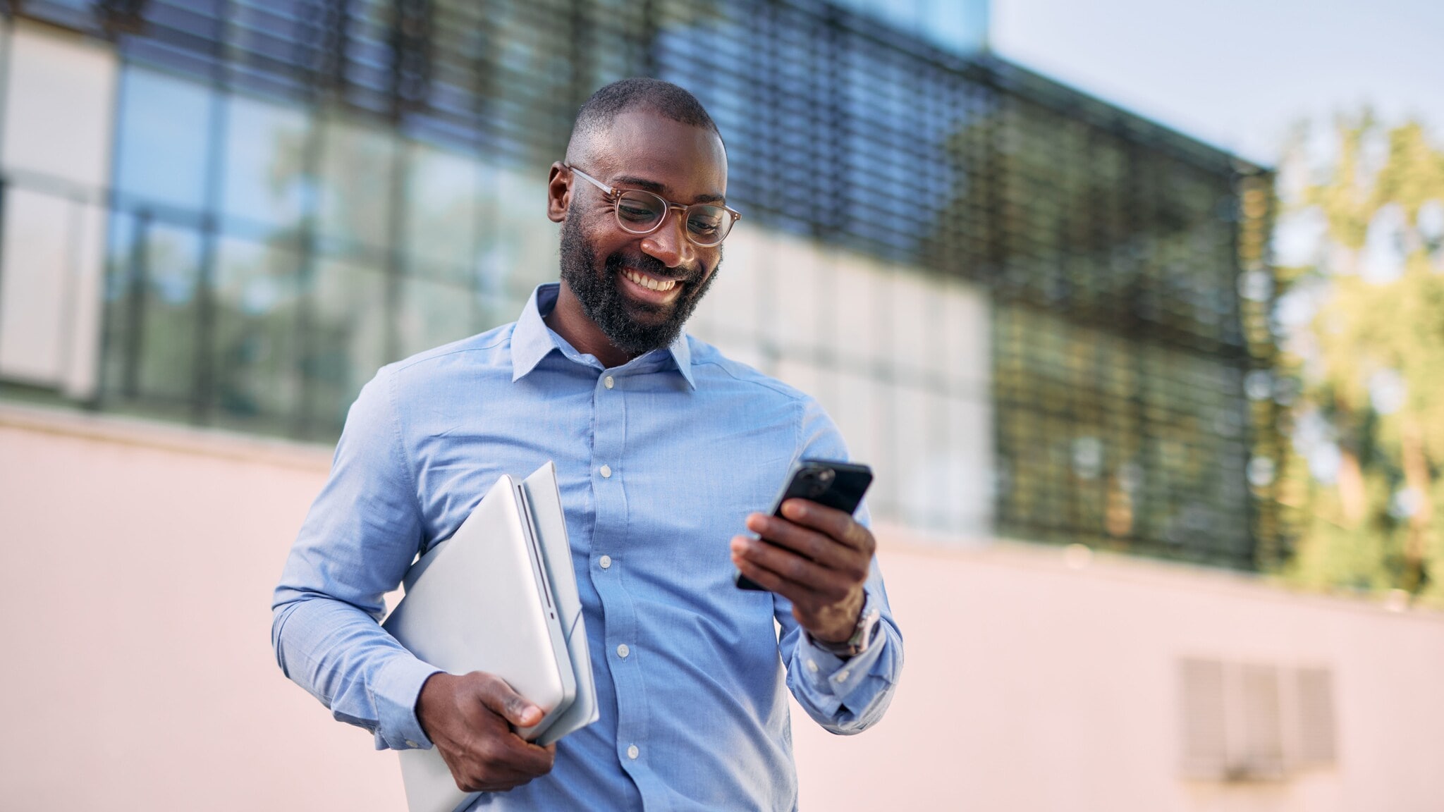 Ein Mann im blauen Hemd mit Laptop unter dem Arm steht vor einem Bürogebäude und blickt erfreut auf ein Smartphone in seiner Hand. Ein Mann im blauen Hemd mit Laptop unter dem Arm steht vor einem Bürogebäude und blickt erfreut auf ein Smartphone in seiner Hand.