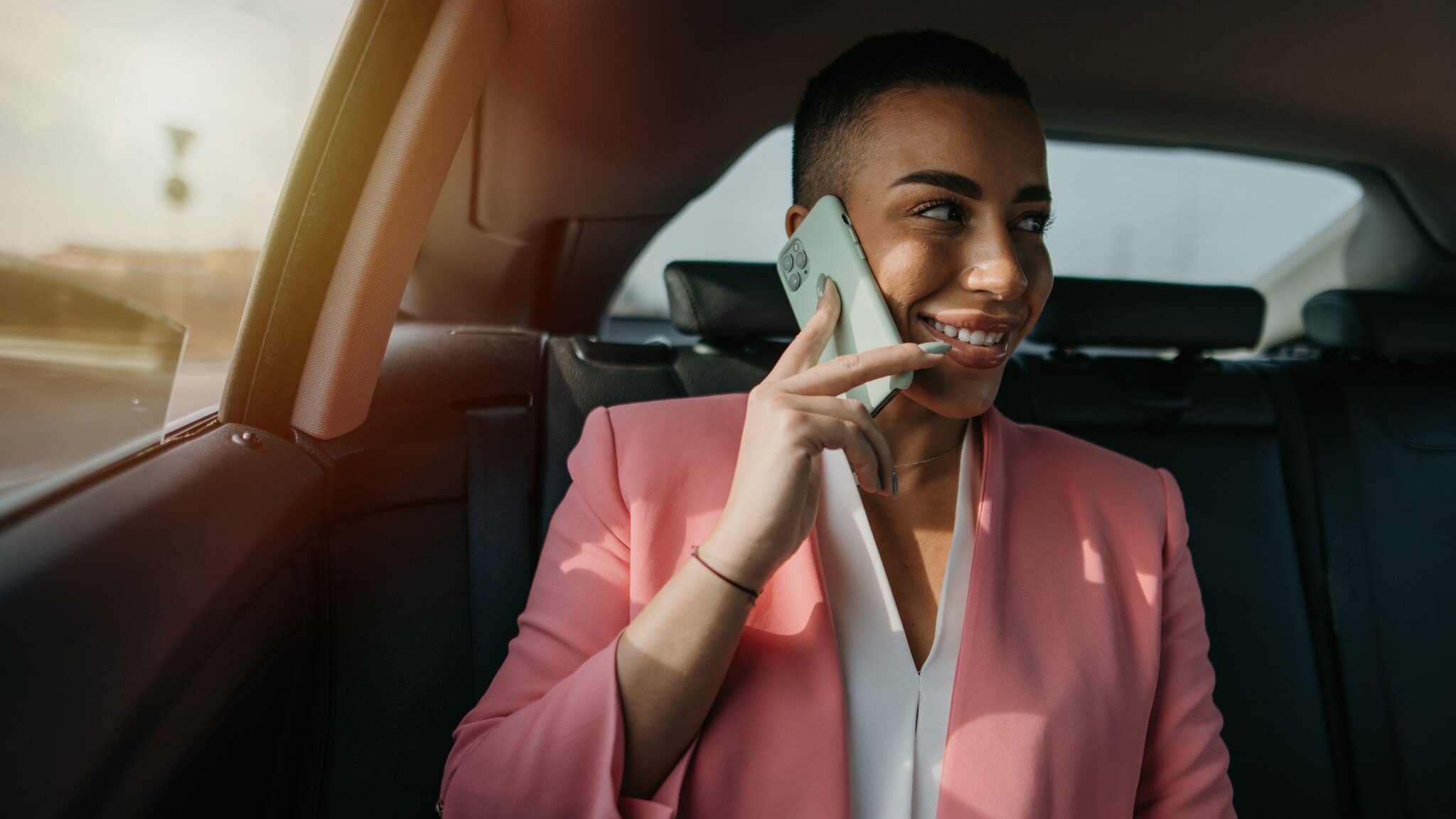 Eine lächelnde Frau im rosafarbenen Blazer sitzt auf der Rückbank eines Autos und telefoniert mit einem Smartphone an ihrem Ohr.