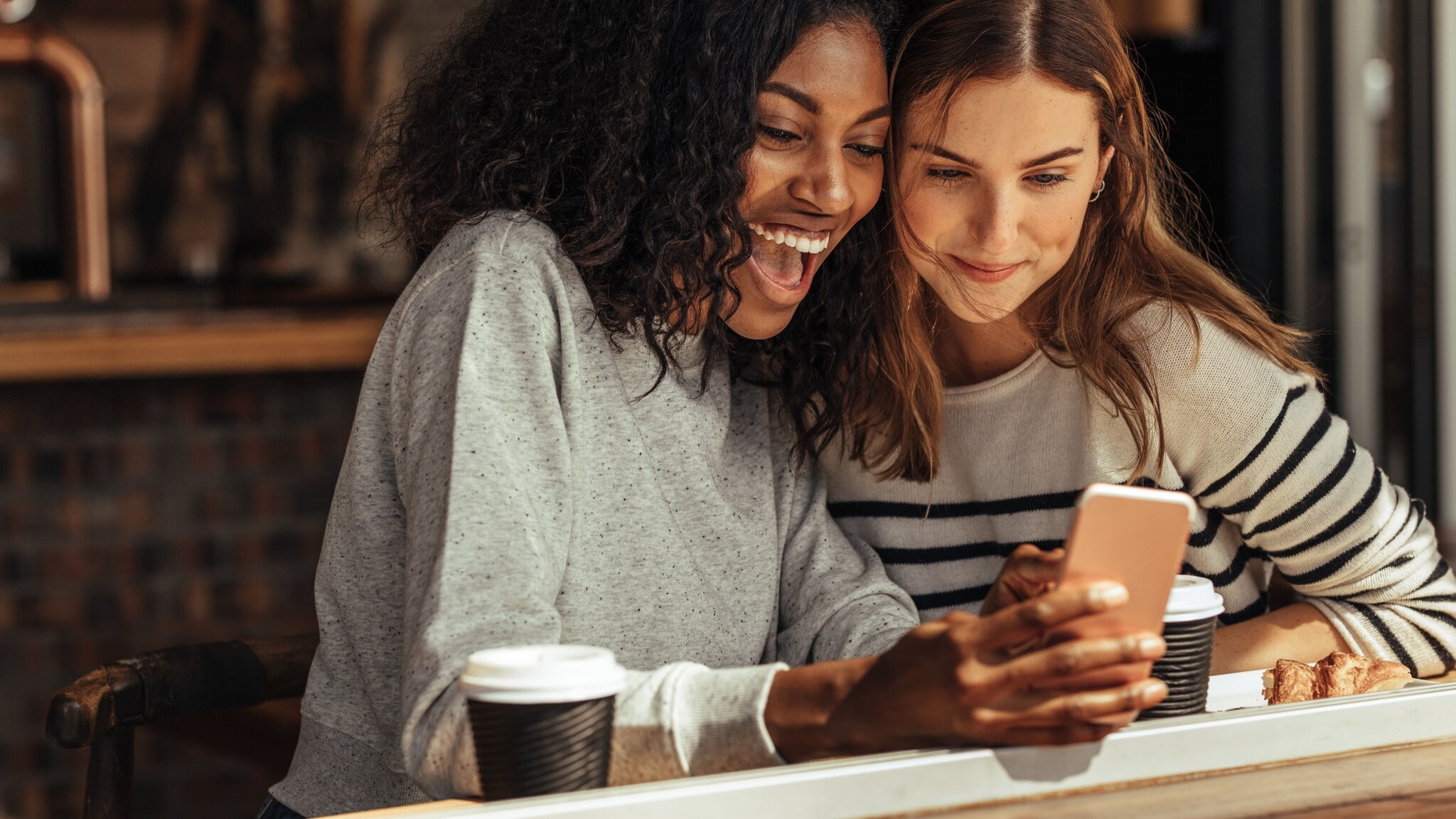 Zwei junge lächelnde Frauen schauen gemeinsam auf ein Smartphone mit Kaffeebechern vor sich. Zwei junge lächelnde Frauen schauen gemeinsam auf ein Smartphone mit Kaffeebechern vor sich.