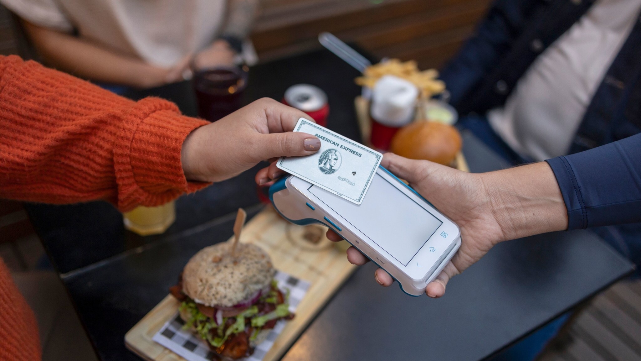 Eine Person hält eine silberne Kreditkarte von American Express an ein Kartenlesegerät, das eine andere Person über einen Tisch mit Hamburgern hält. Eine Person hält eine silberne Kreditkarte von American Express an ein Kartenlesegerät, das eine andere Person über einen Tisch mit Hamburgern hält.