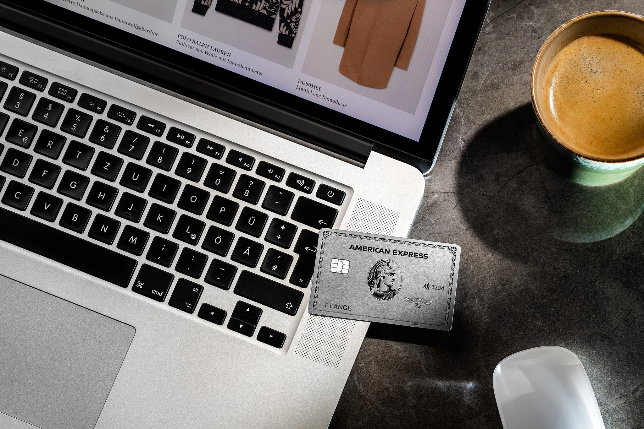 Aufsicht einer silbernen Kreditkarte von American Express auf einem Laptop neben einer Tasse Kaffee auf einem Tisch.