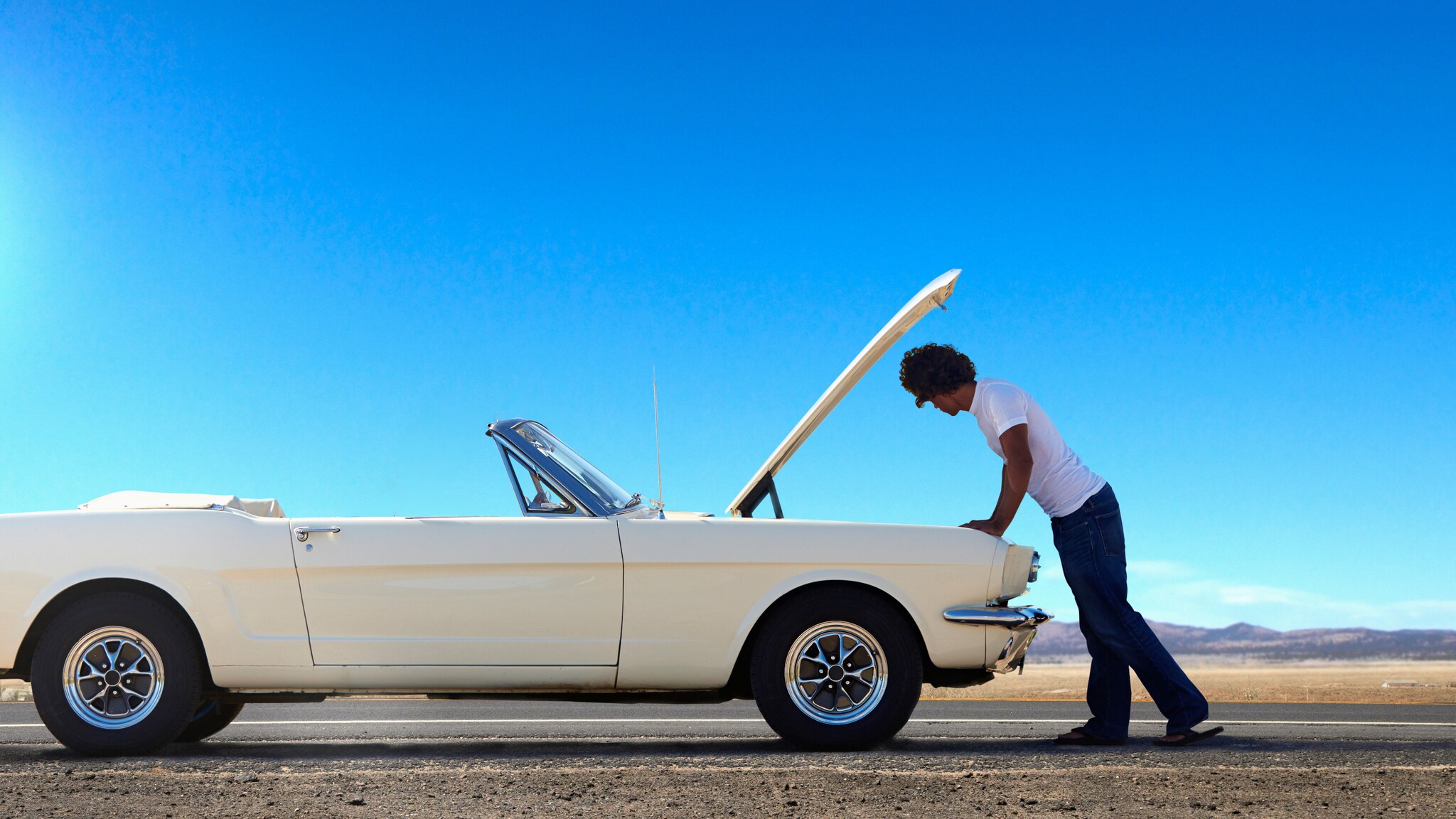 Ein Mann in weißem T-Shirt inspiziert unter der geöffneten Kühlerhaube den Motorraum eines weißen Cabriolets auf einer Straße in einer Wüstenlandschaft.