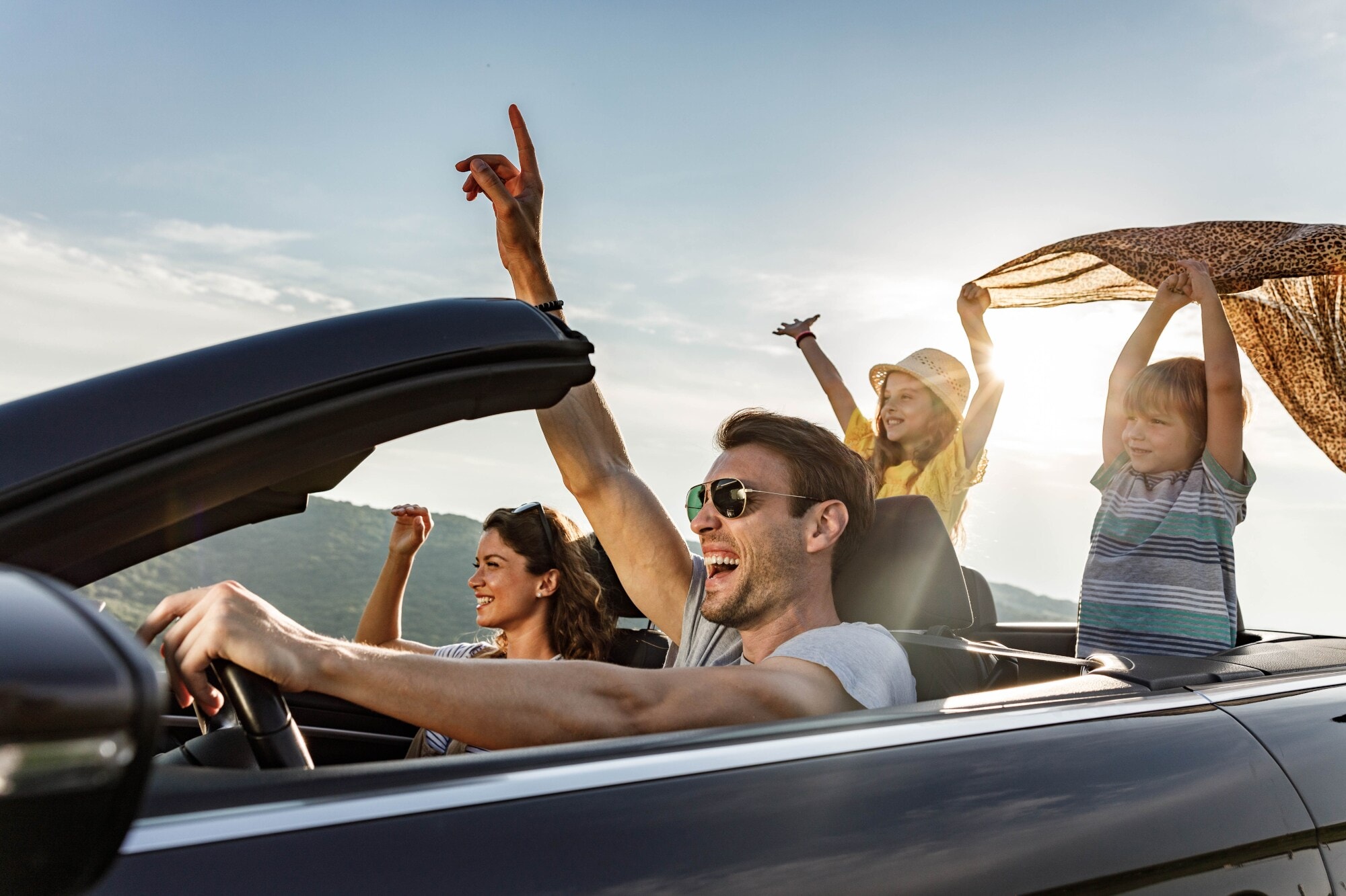 Eine Familie mit zwei Kindern fährt mit ausgestreckten Händen in einem Cabriolet bei Sonnenschein. Eine Familie mit zwei Kindern fährt mit ausgestreckten Händen in einem Cabriolet bei Sonnenschein.