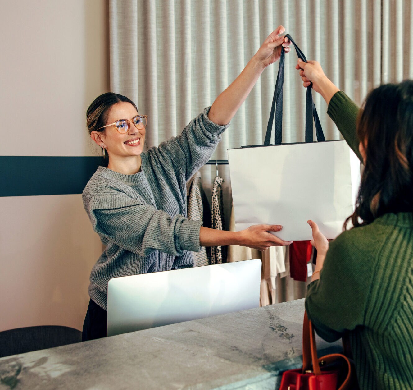 Eine lächelnde Verkäuferin reicht einer Kundin eine Einkaufstasche über den Kassentresen in einer schicken Modeboutique