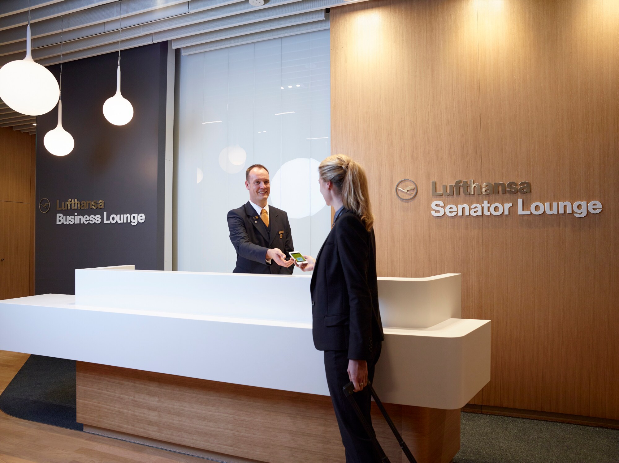 Eine Frau checkt bei einem Mitarbeiter an der Rezeption einer Lufthansa Lounge ein. Eine Frau checkt bei einem Mitarbeiter an der Rezeption einer Lufthansa Lounge ein.