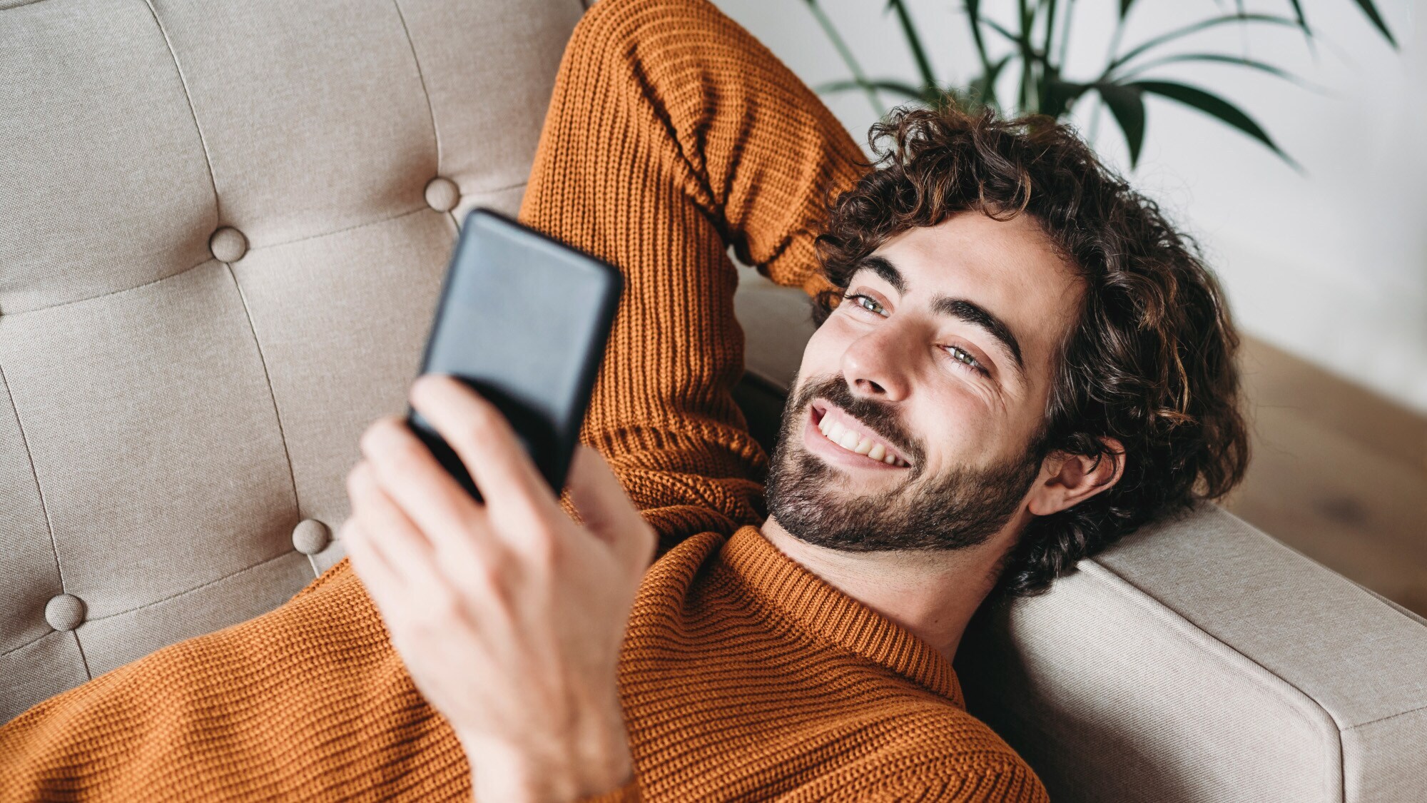 Ein junger Mann liegt entspannt auf dem Sofa und schaut lächelnd auf sein Smartphone.