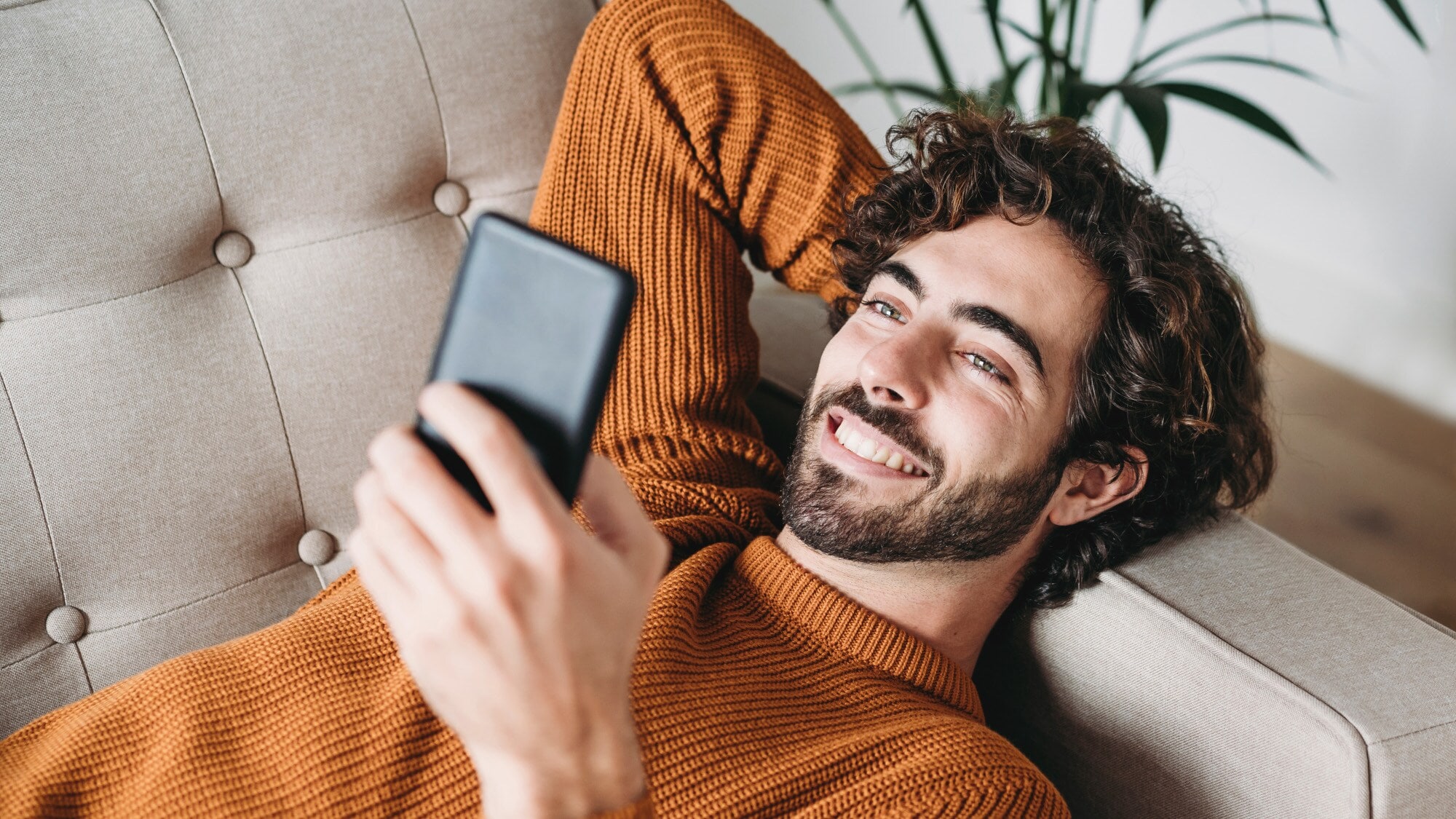 Ein junger Mann liegt entspannt auf dem Sofa und schaut lächelnd auf sein Smartphone. Ein junger Mann liegt entspannt auf dem Sofa und schaut lächelnd auf sein Smartphone.