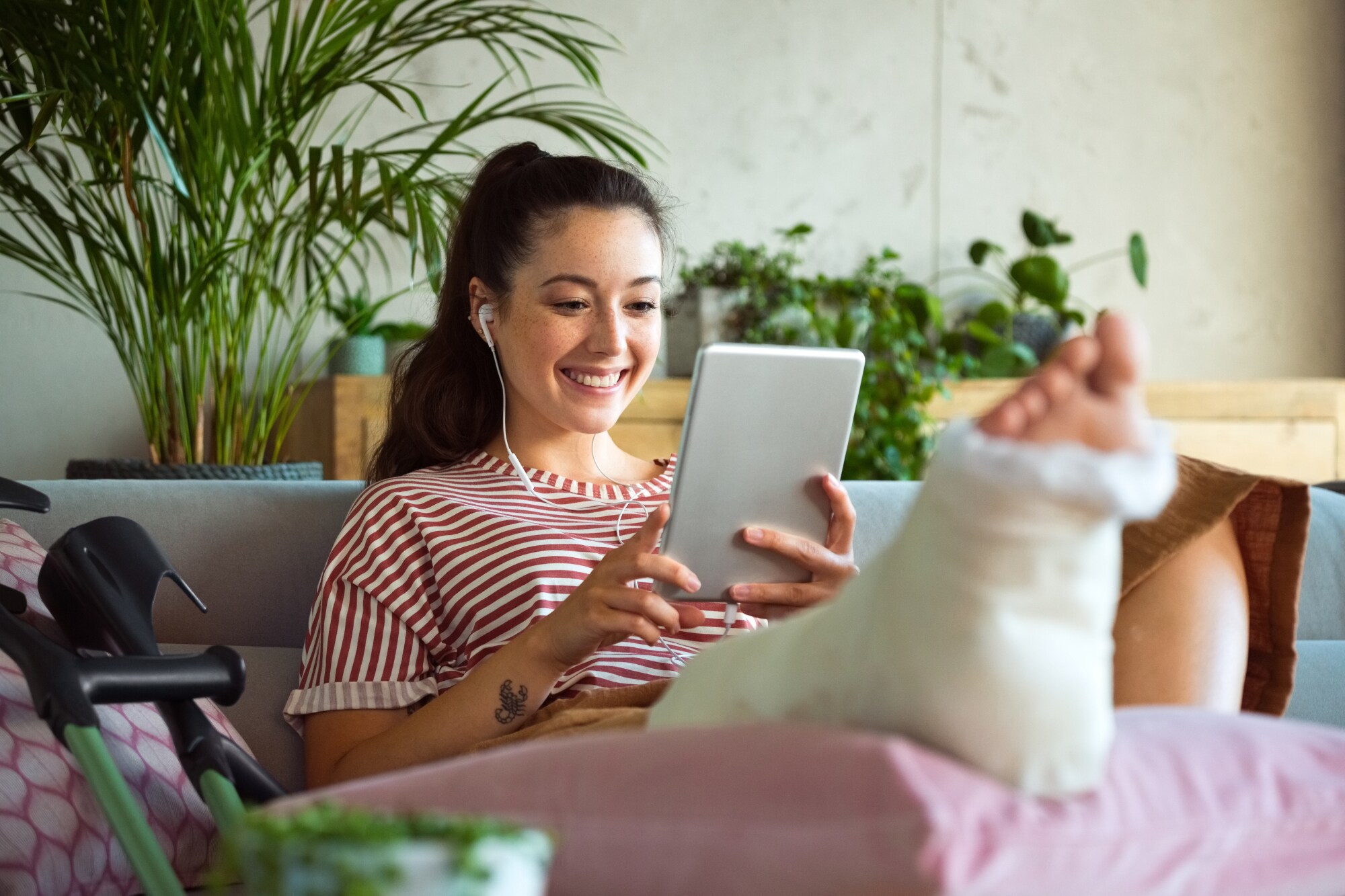 Eine junge Frau auf einem Sofa schaut lächelnd auf ihr Tablet, während sie ihr eingegipstes Bein hochgelegt hat