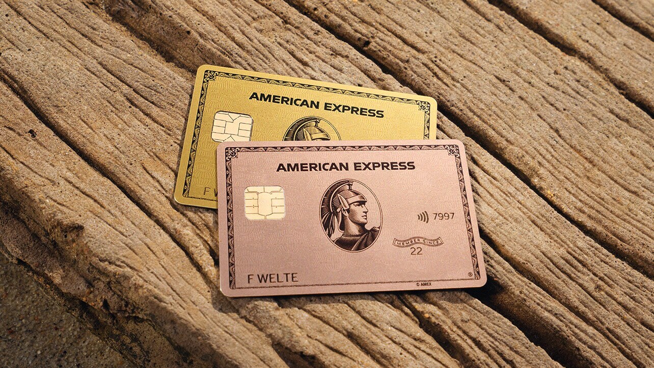 Eine goldene und eine rosafarbene American Express Kreditkarte aus Metall liegen auf einem Stück Holz im Sonnenlicht.