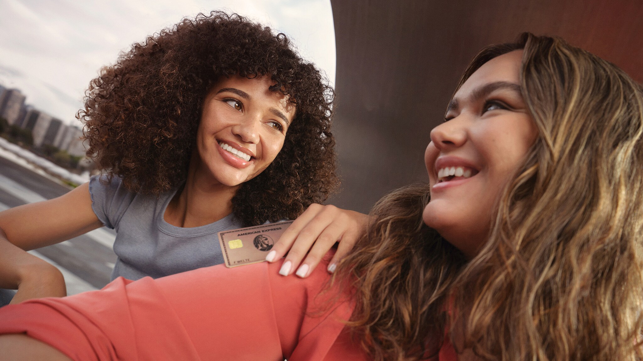 Zwei junge Frauen schauen sich lächelnd an, eine hat die Gold Card Rosé aus Metall von American Express in der Hand.