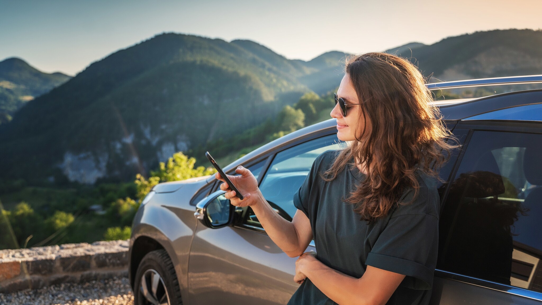 Eine junge Frau mit Sonnenbrille lehnt an der Seite eines Autos und blickt auf ihr Smartphone, im Hintergrund grüne Berglandschaft bei Sonnenschein. Eine junge Frau mit Sonnenbrille lehnt an der Seite eines Autos und blickt auf ihr Smartphone, im Hintergrund grüne Berglandschaft bei Sonnenschein.